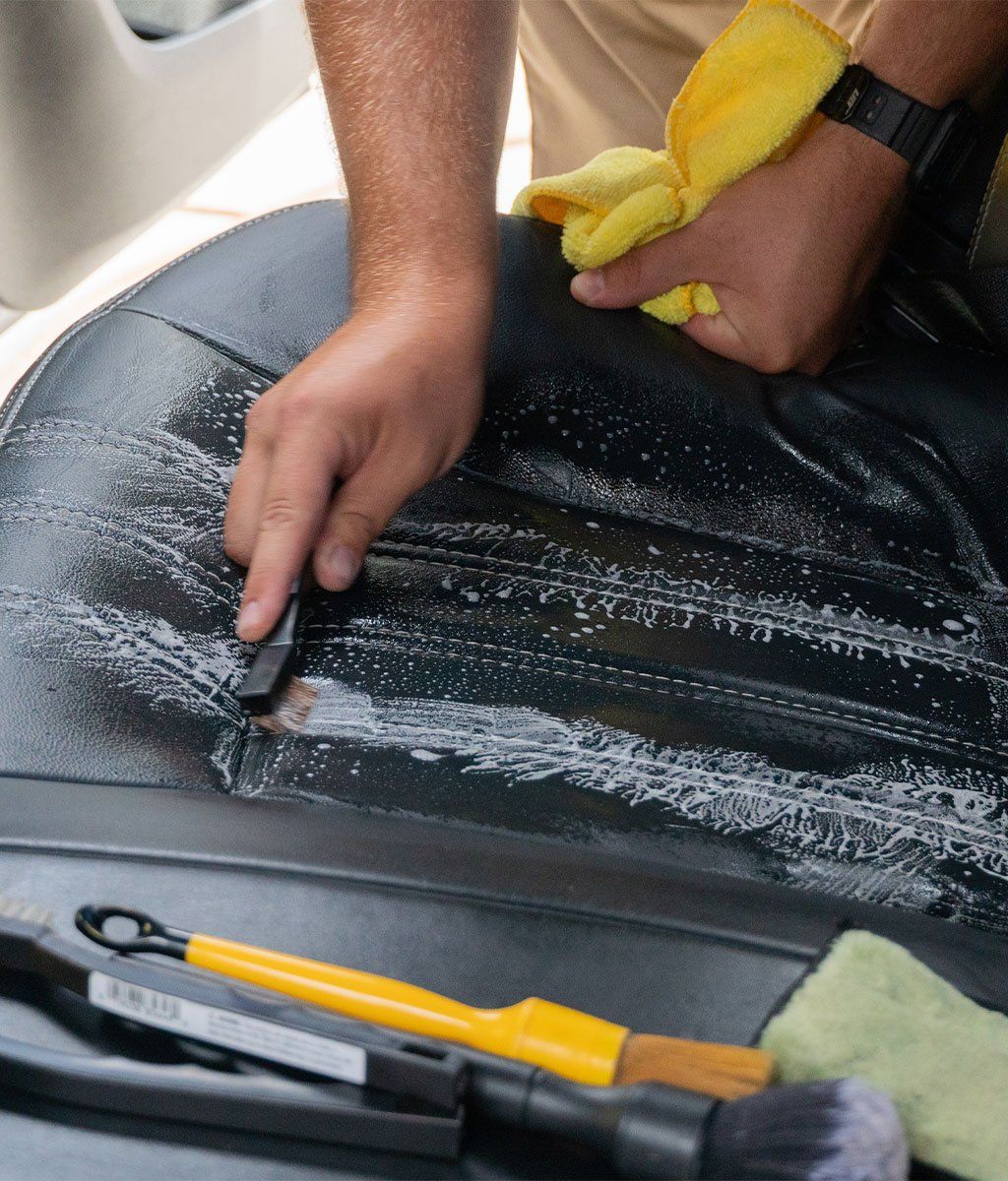 Person cleaning a black car seat with a sponge and scraper, with soap suds visible