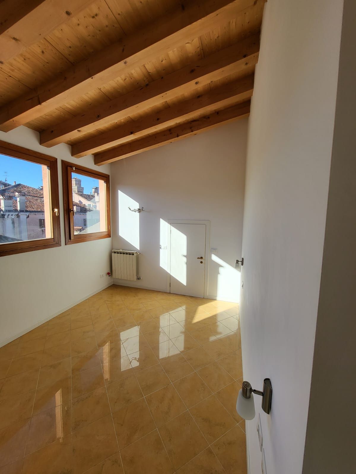 Stanza vuota e luminosa con travi a vista sul soffitto, pavimento in parquet e ampie finestre.