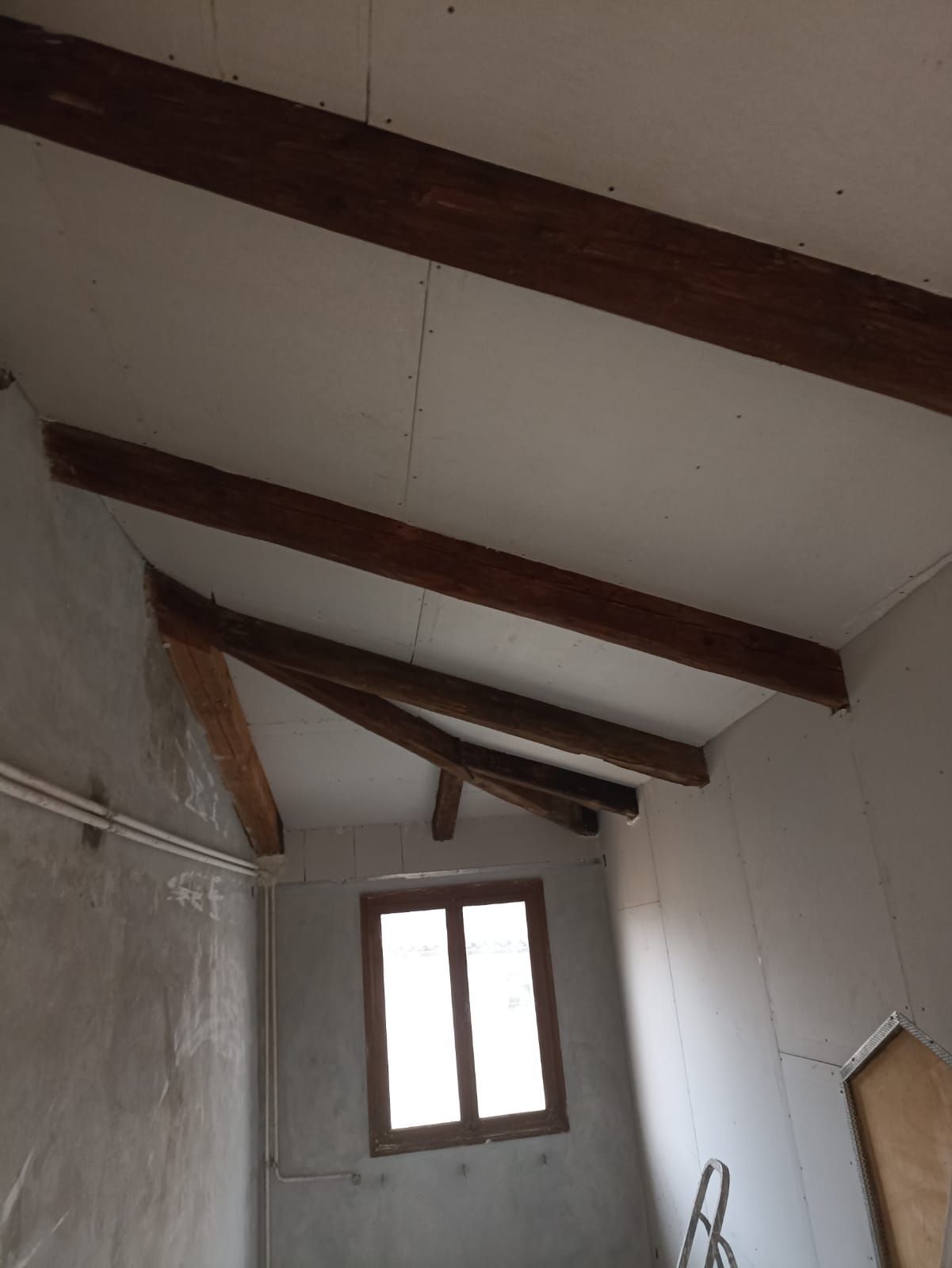 Stanza vuota con soffitto bianco inclinato, travi scure a vista e una piccola finestra.