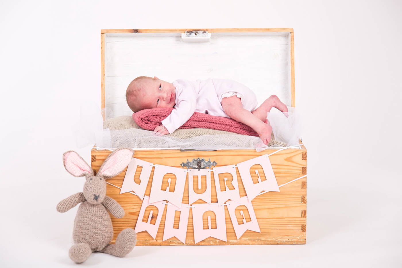 Neugeborenes Baby in einer Holztruhe mit rosa Decke, Hasenspielzeug und Banner mit der Aufschrift