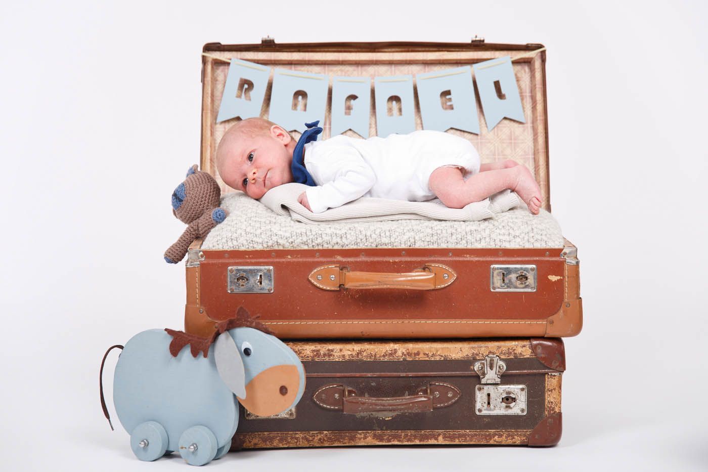 Baby liegt auf gestapelten Vintage-Koffer, blaues Namensbanner, Spielzeugesel davor.