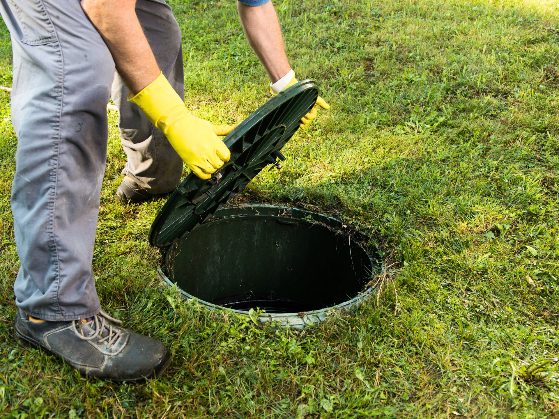 A man wearing yellow gloves is opening a manhole cover in the grass.