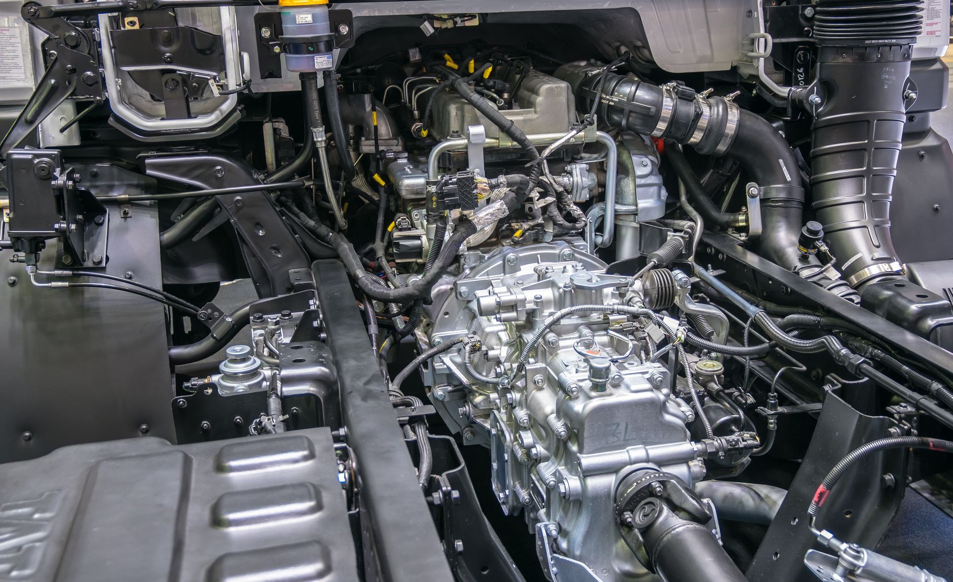 Car engine bay with exposed silver transmission and black hoses in a workshop setting