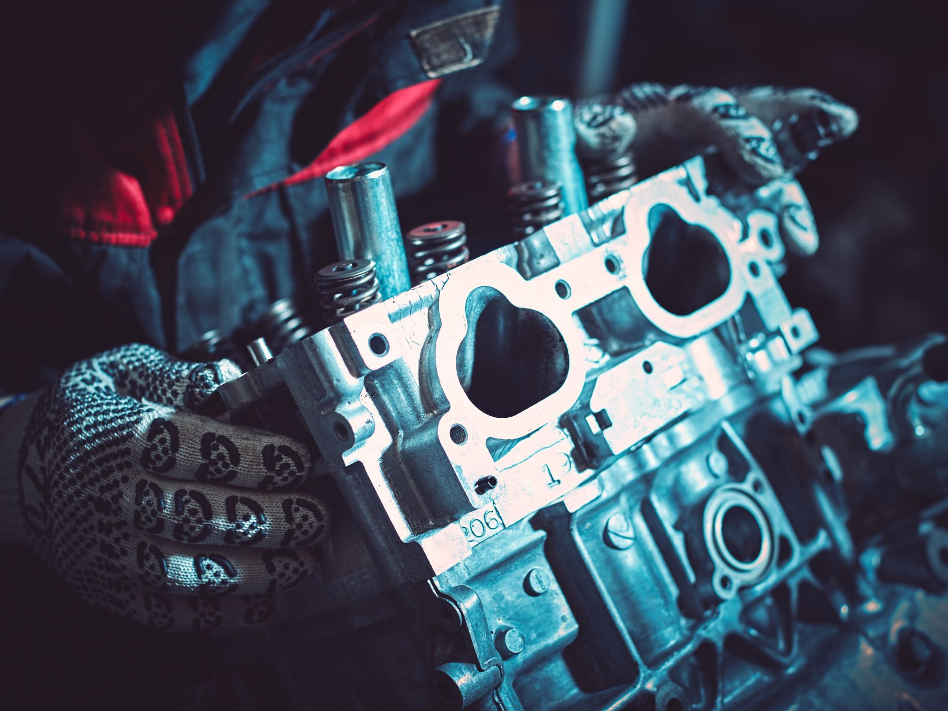 Mechanic's gloved hands holding a car engine cylinder head in a dim, blue-lit workshop