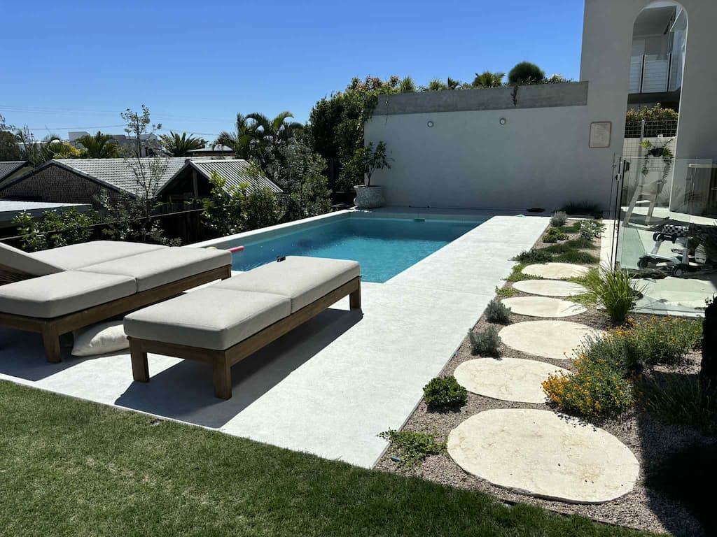 Modern backyard pool with white deck, two loungers, and stone stepping path beside greenery.