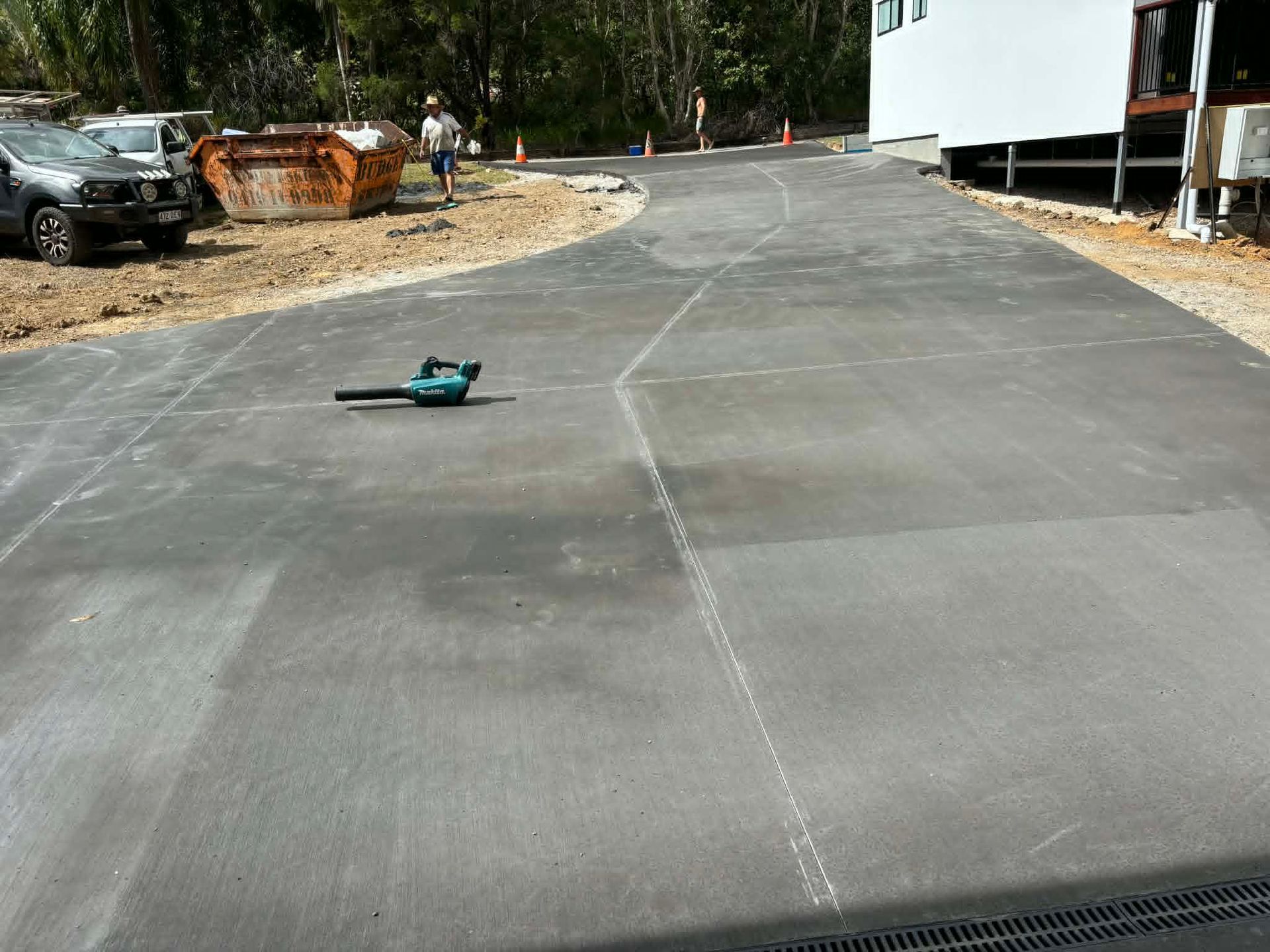 A green power tool lies on a paved driveway near parked vehicles and a white building.