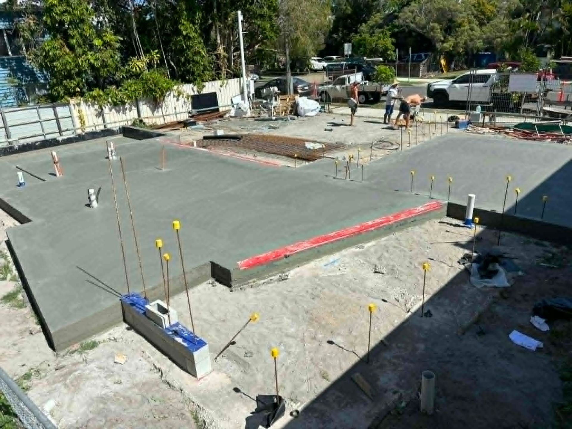 Concrete foundation slab under construction with workers, rebar, and yellow markers in a sunny lot