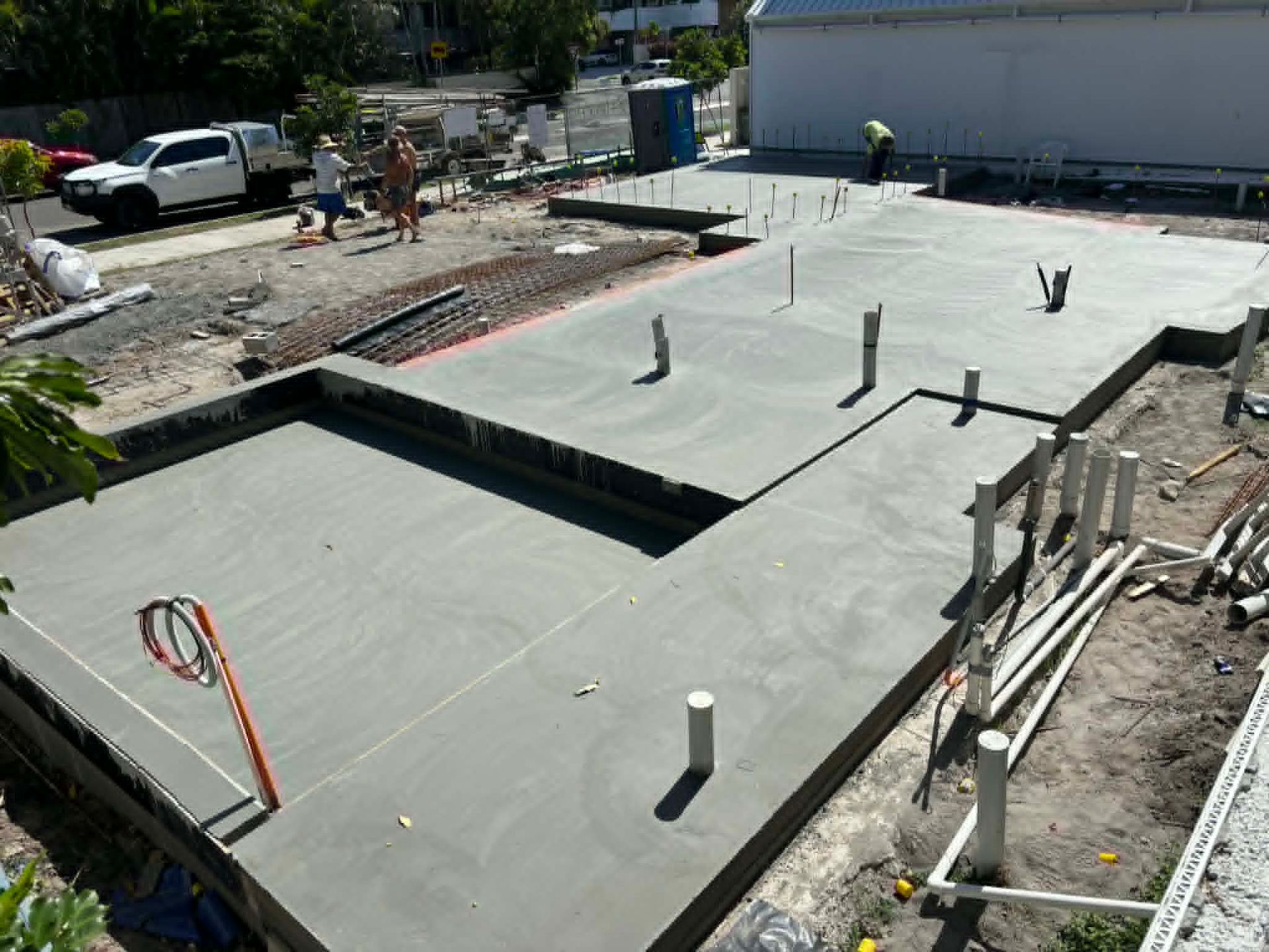 Concrete foundation under construction with exposed pipes and workers in the background