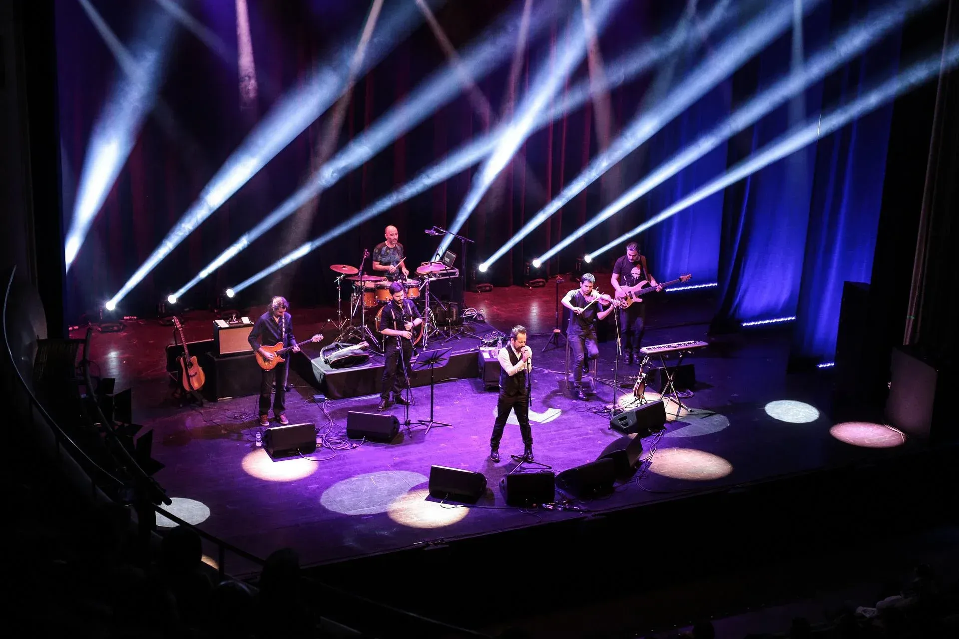 Band on stage with bright spotlights; musicians playing instruments; vocalist singing.
