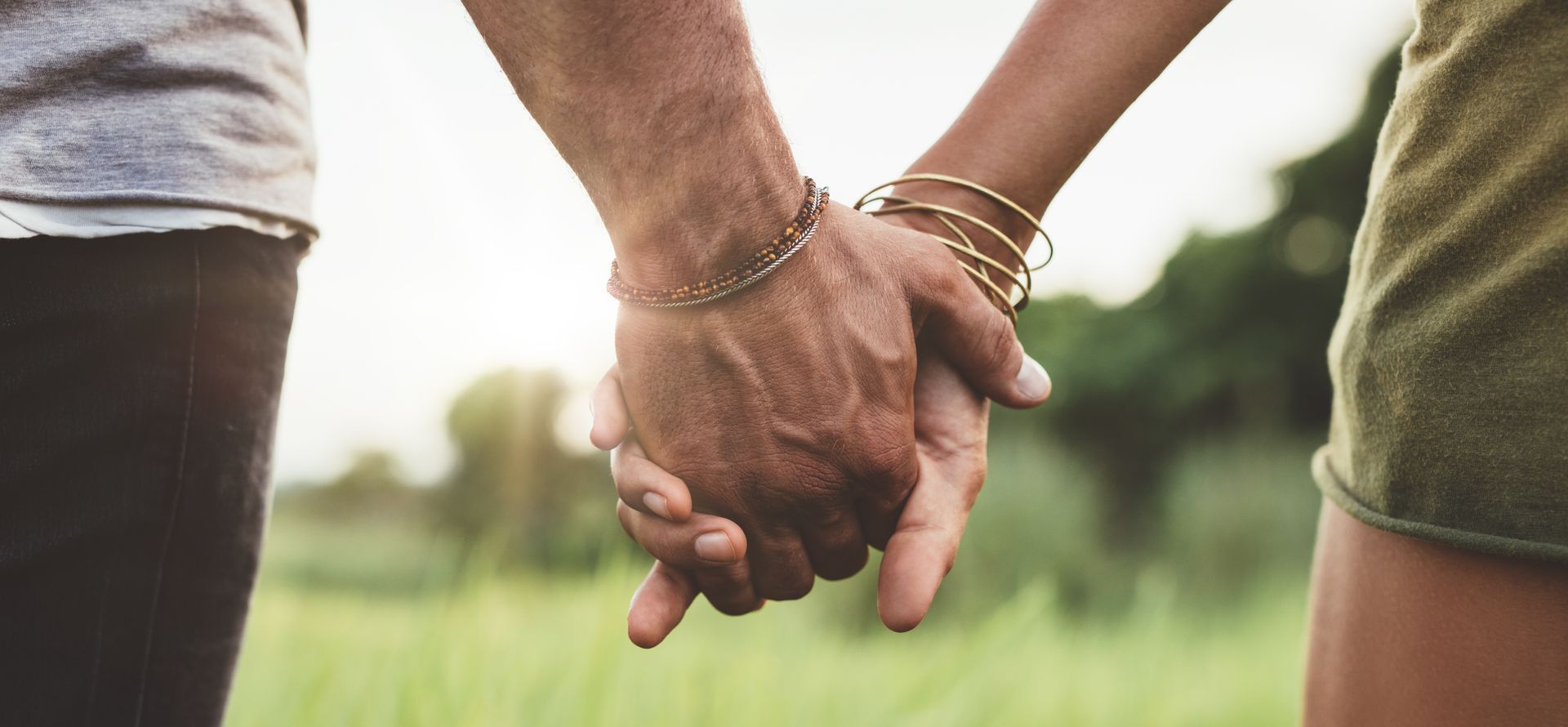 A couple holding hands outdoors, with a grassy background. Focus on the clasped hands.