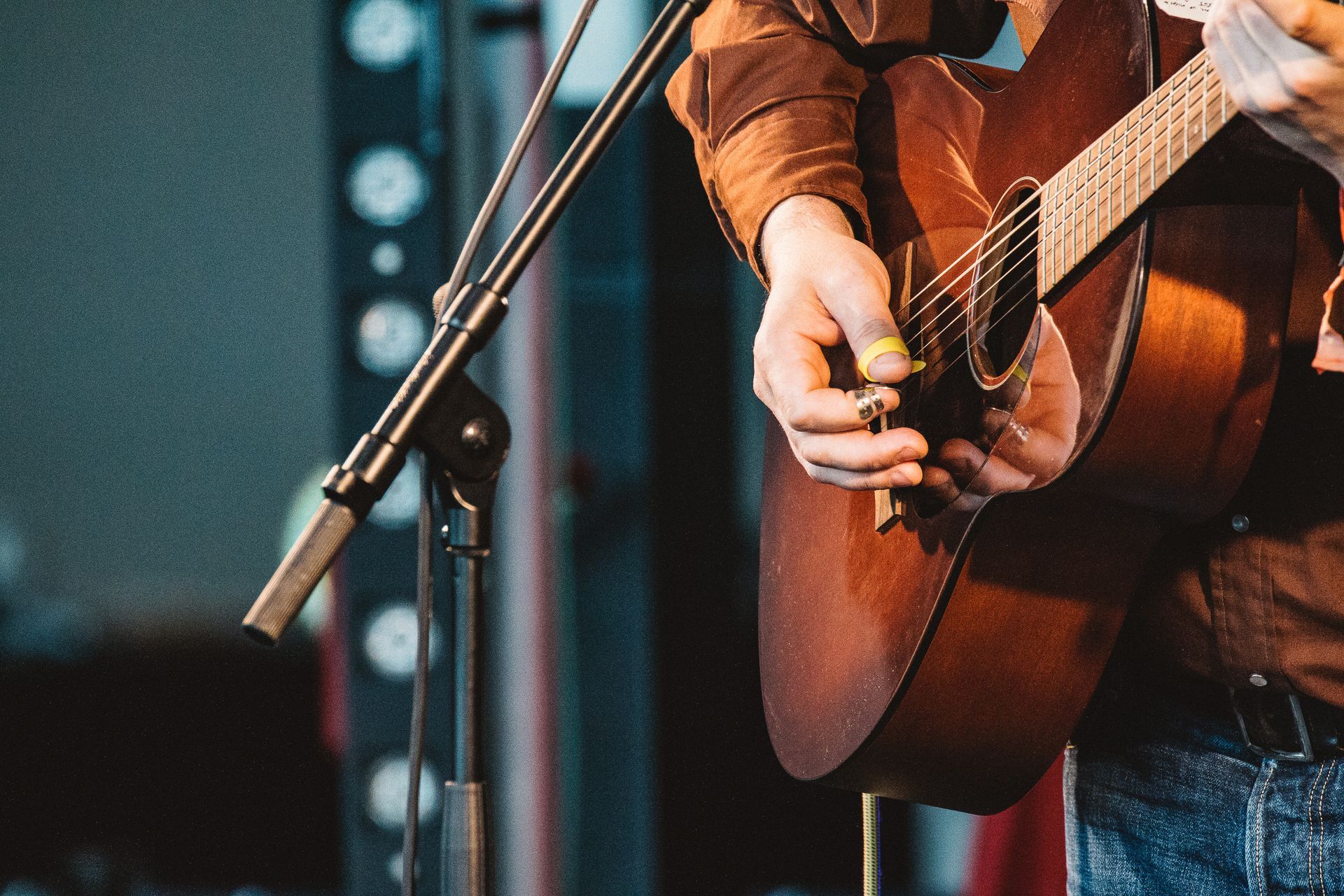 Person playing an acoustic guitar on stage, strumming strings with a pick near a microphone.