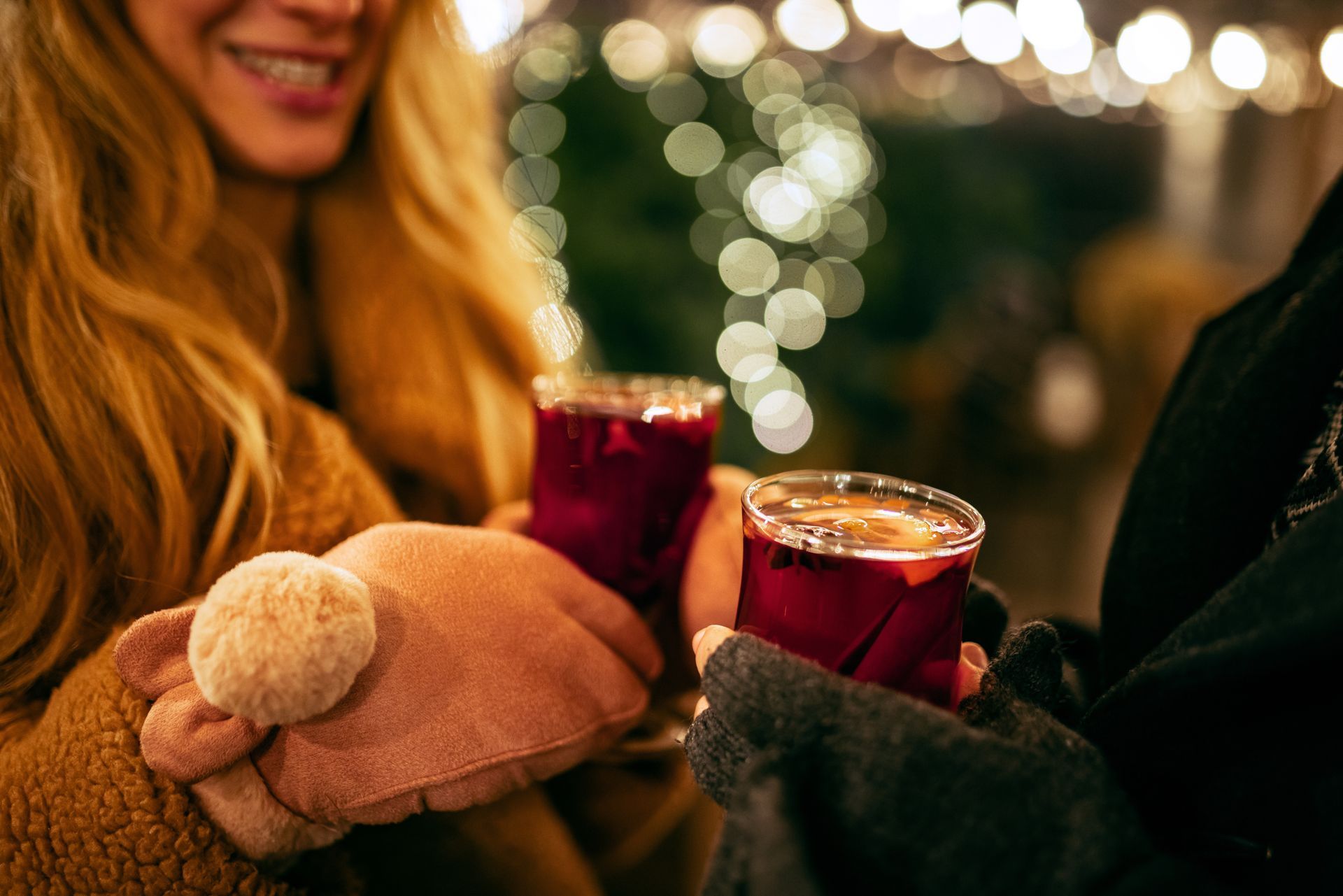 People toasting mulled wine at a winter market. One wears pink mittens, blurred lights in the background.