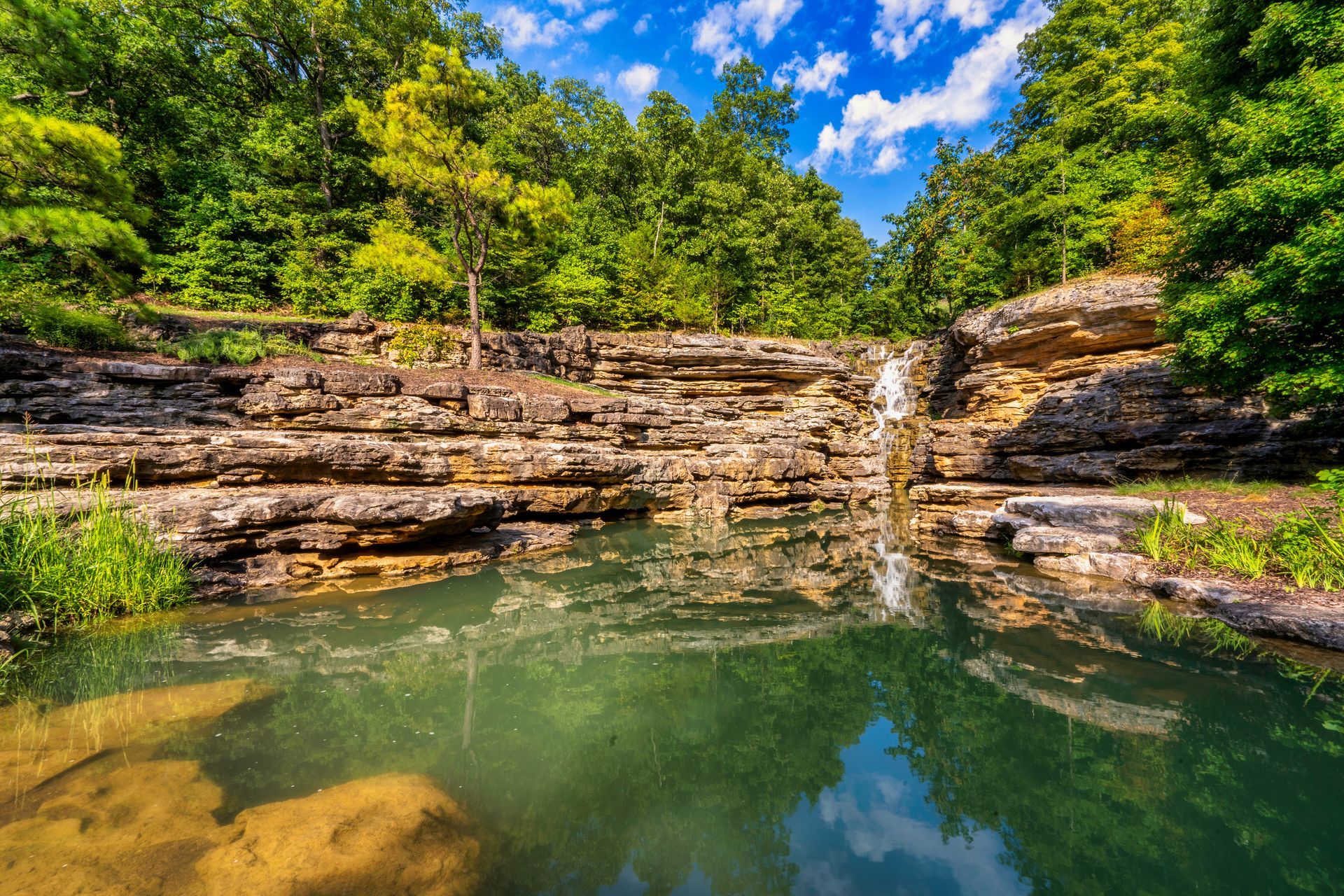 Waterfall cascading into a clear pool. Brown rock ledge with lush green trees, blue sky with clouds.