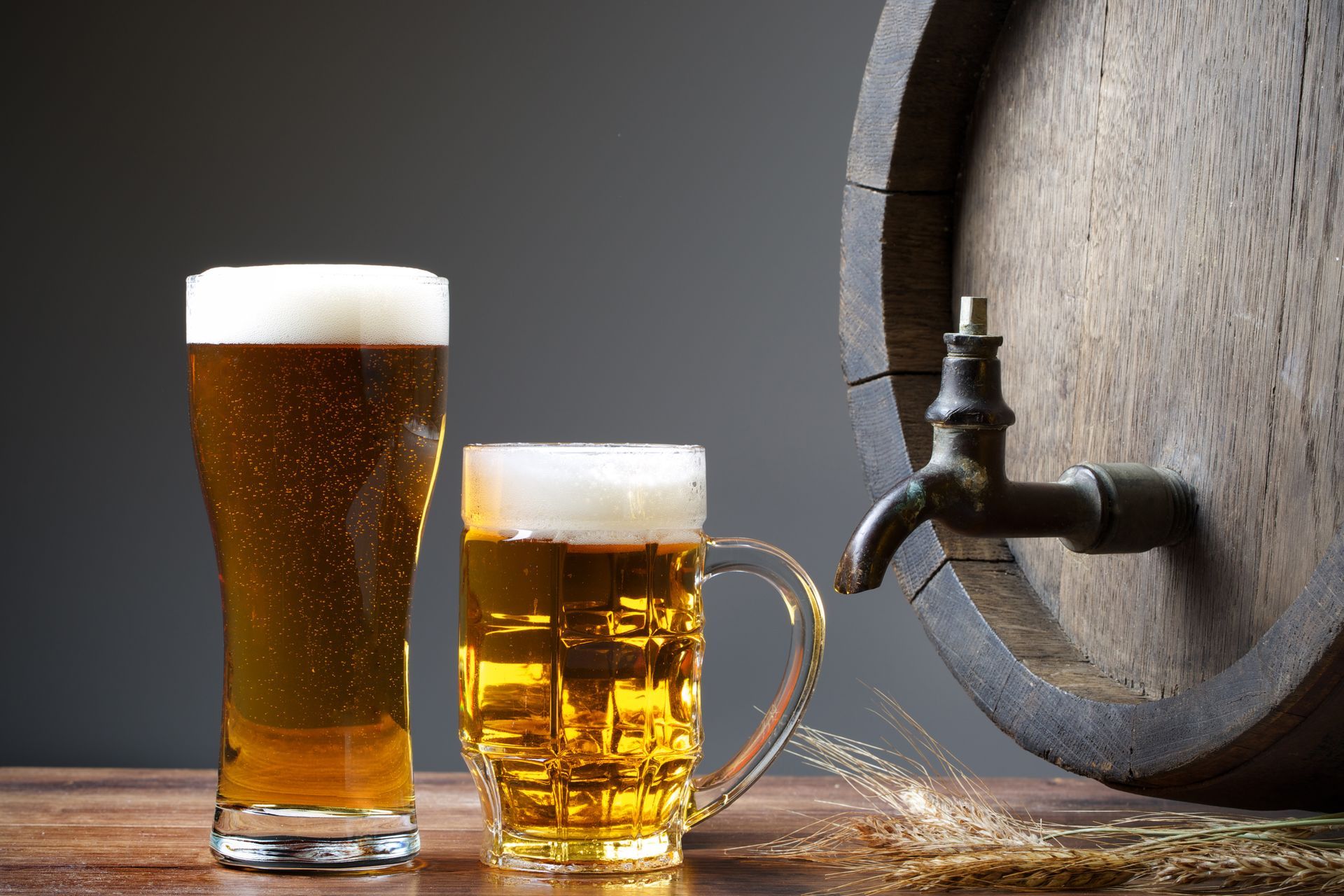 Two glasses of beer with foamy heads, near a wooden barrel with tap.
