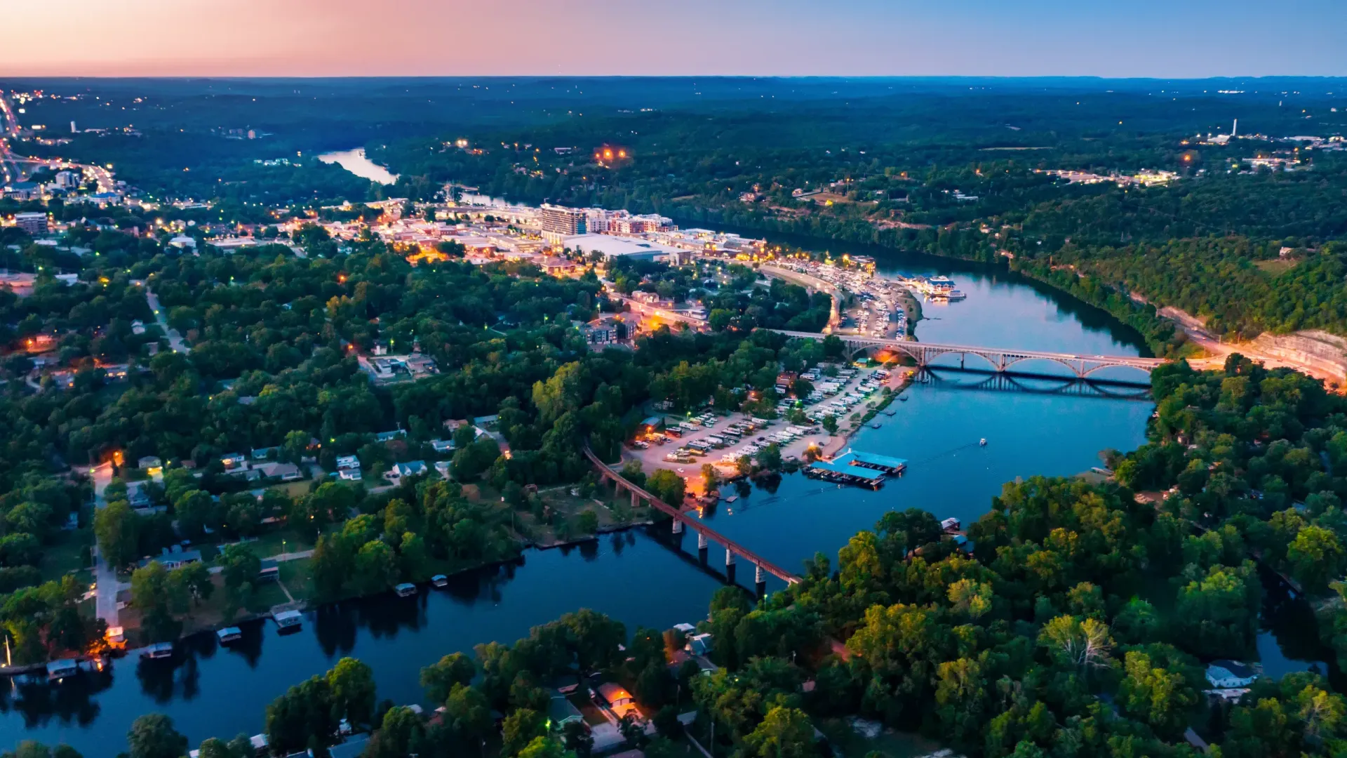 Aerial view of a brightly lit city nestled along a dark river at dusk, surrounded by lush green trees.