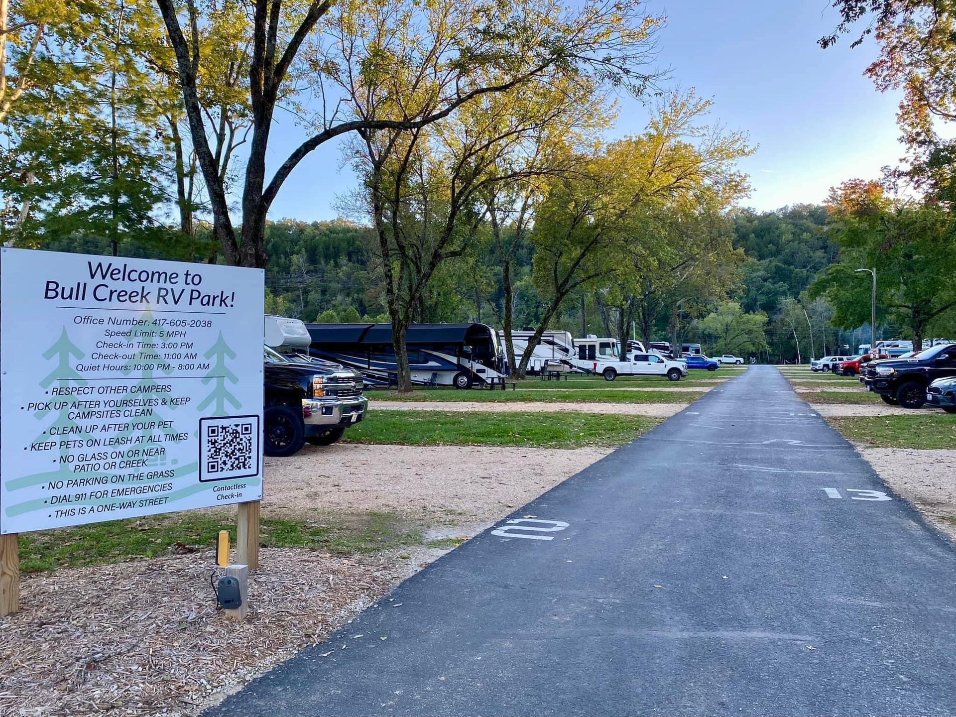 RV park entrance with sign, vehicles parked along road, trees in background.