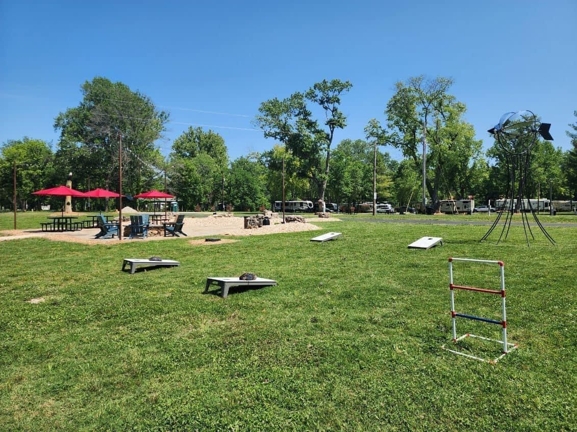 Lush green park with cornhole boards and ladder toss set up. People are in the background under shade trees.