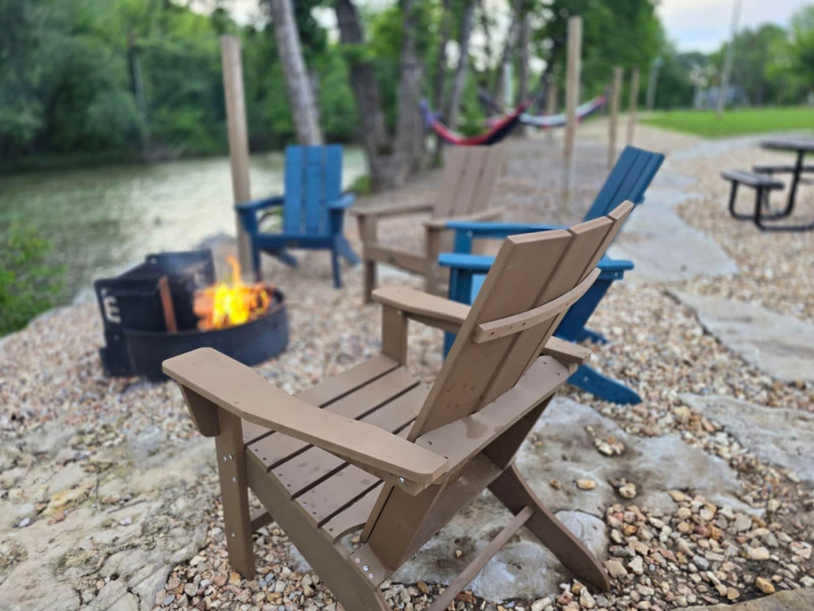 Chairs around a fire pit by a river; two blue chairs, one tan. Hammock and picnic table in background.