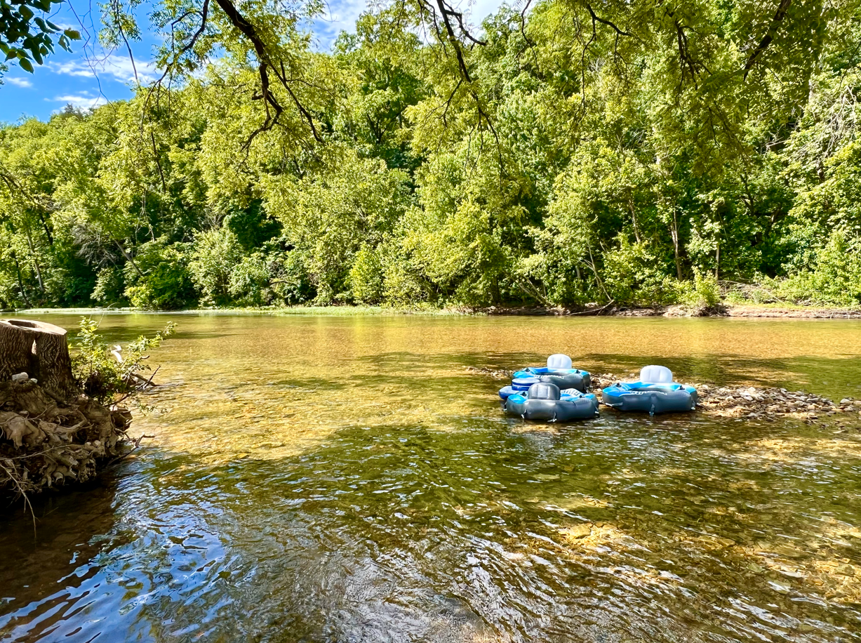 Two blue river tubes in a shallow, clear river; green trees line the shore under a sunny sky.