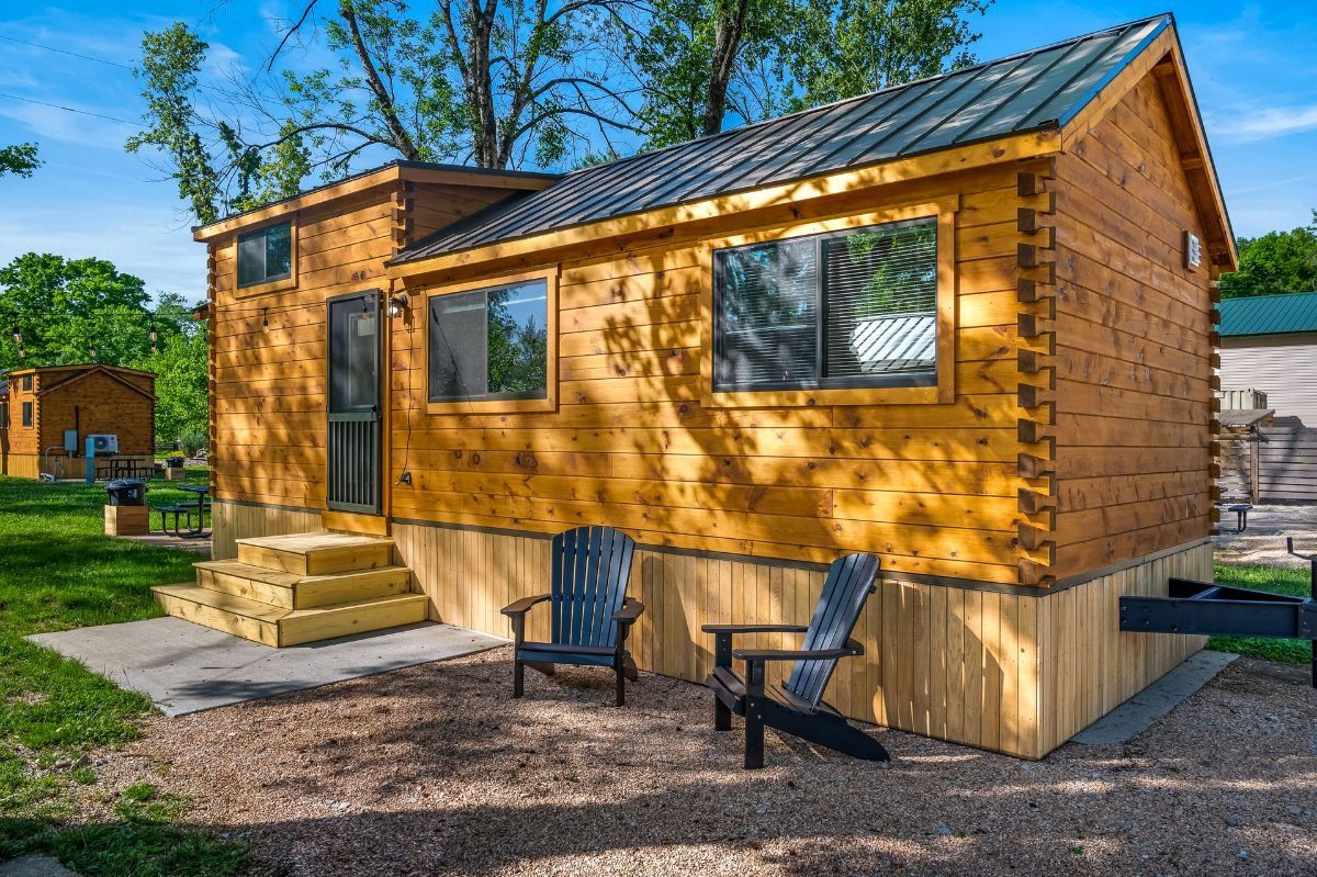 Small wooden cabin with a dark gray metal roof, two chairs in front, and a grassy yard.