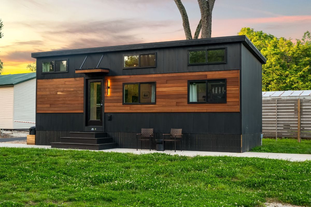 Two-story tiny house with black and wood siding, small steps, and chairs outside on the grass.