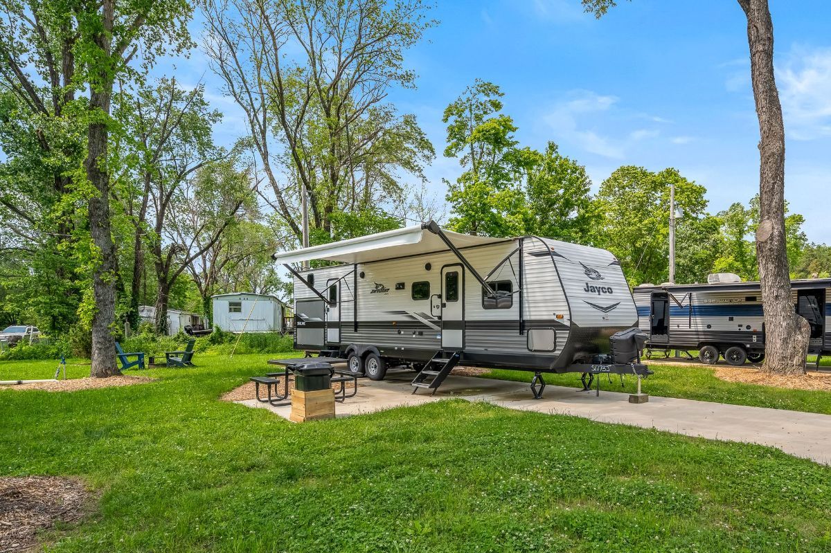 RV parked at a campsite, green grass, trees, and a blue sky.