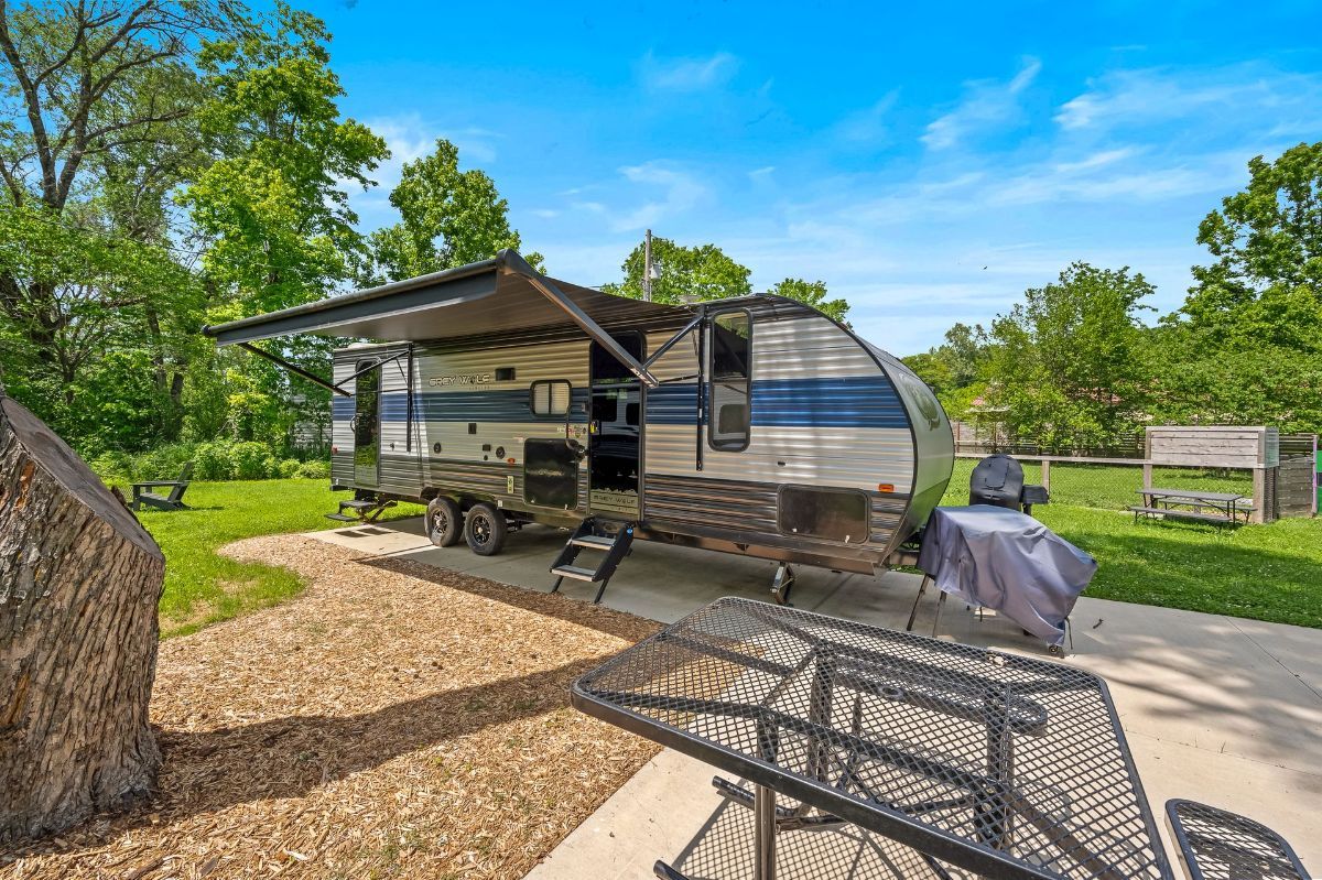 RV parked on a gravel and concrete pad with awning open, blue and silver color, sunny day.