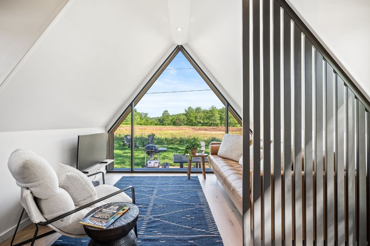 Cozy attic sitting area with a large triangular window overlooking a field, white walls, and a blue rug.