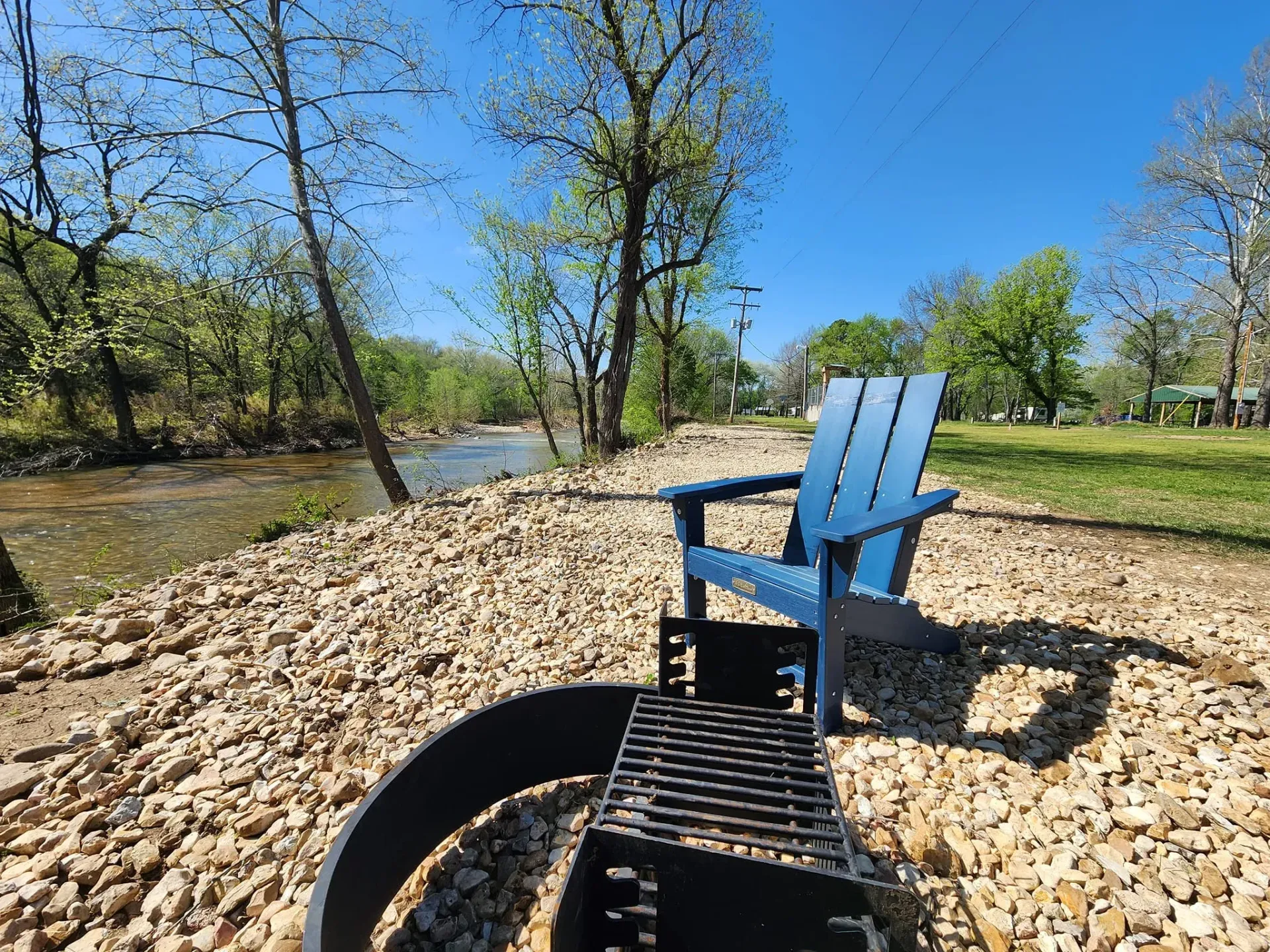 Blue Adirondack chair by a river and fire pit on a bed of rocks, sunny day.