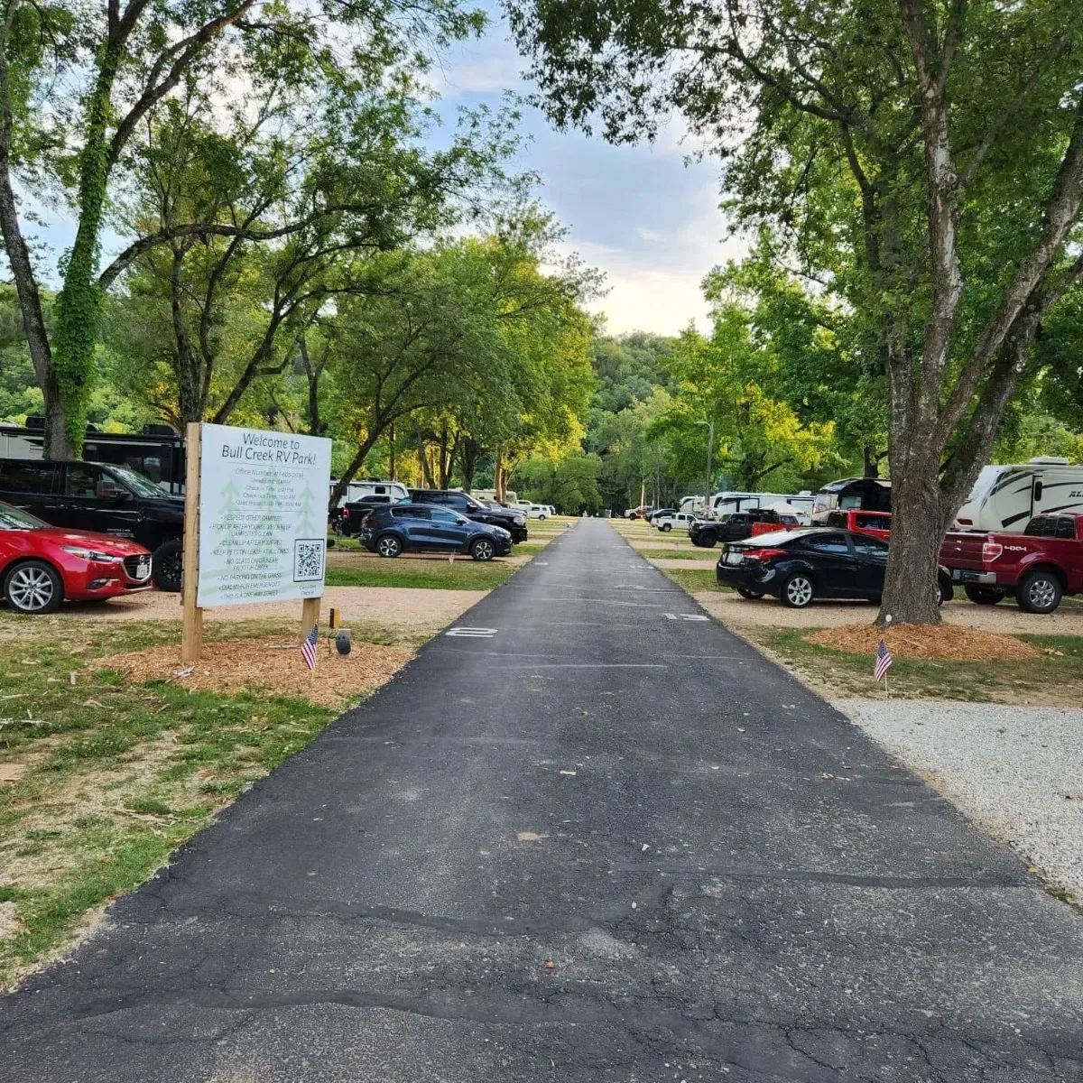 Paved road through a campground lined with RVs, cars, and trees on a sunny day.