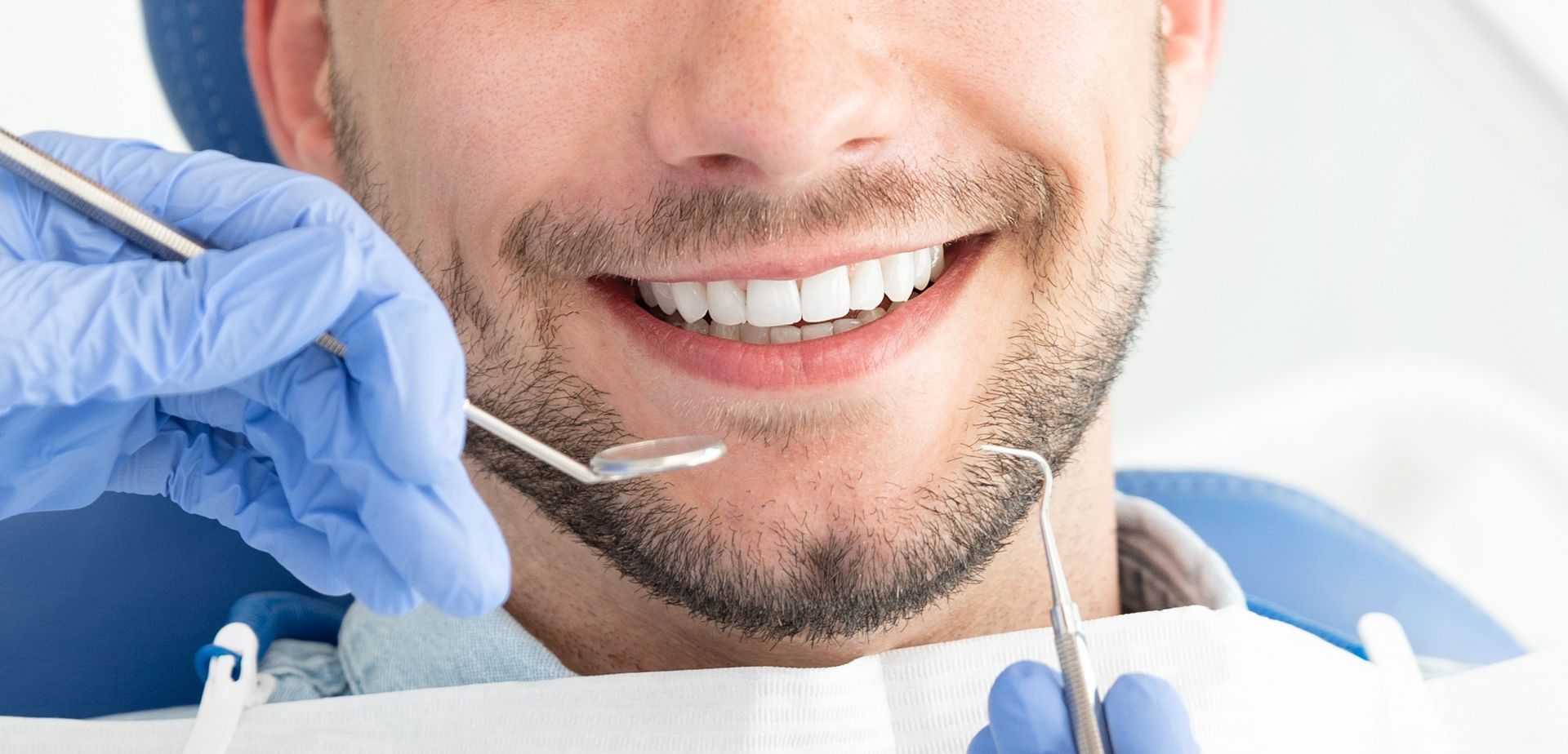 Dentist examining a smiling patient’s teeth with dental tools and blue gloves