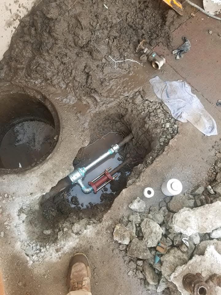 Trench dug in concrete revealing water pipe and valve, near a manhole, ground.