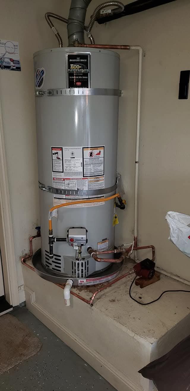 A water heater in a utility room, gray tank with connected pipes, resting on a concrete platform.