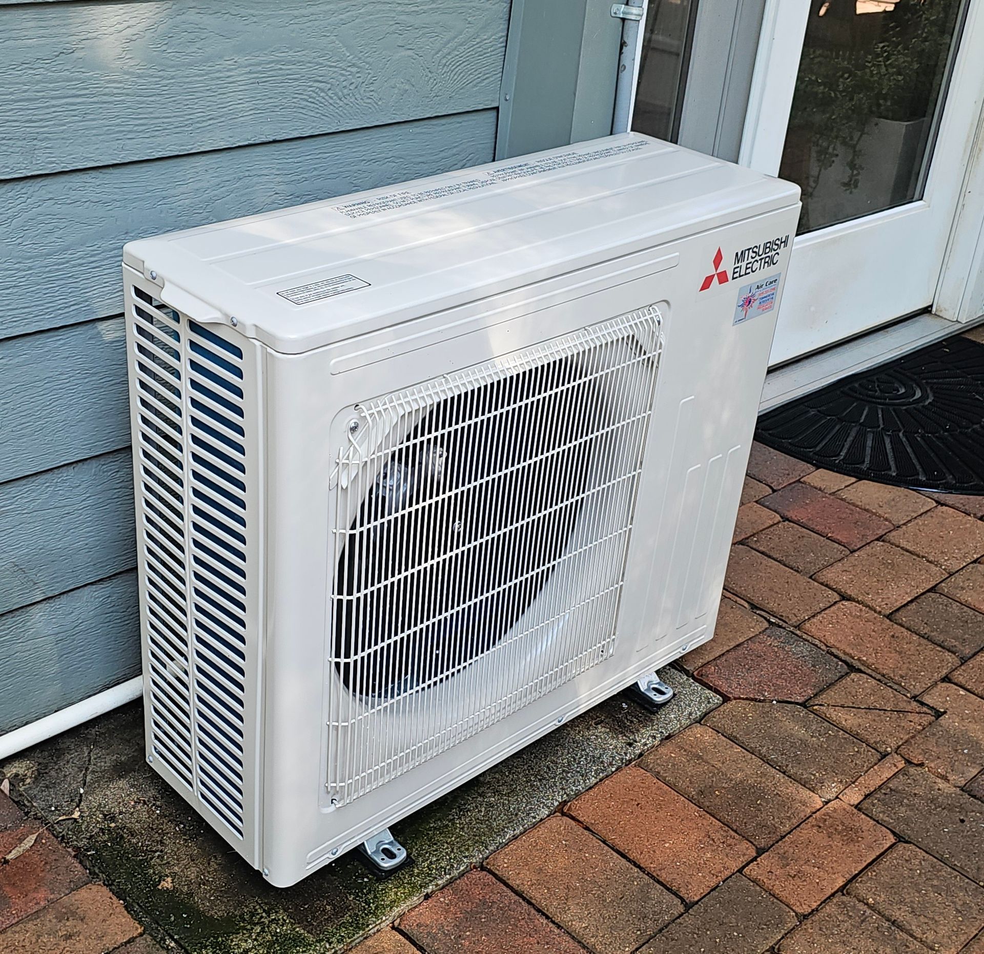 Outdoor air conditioner unit beside a house wall and white door on a brick patio.