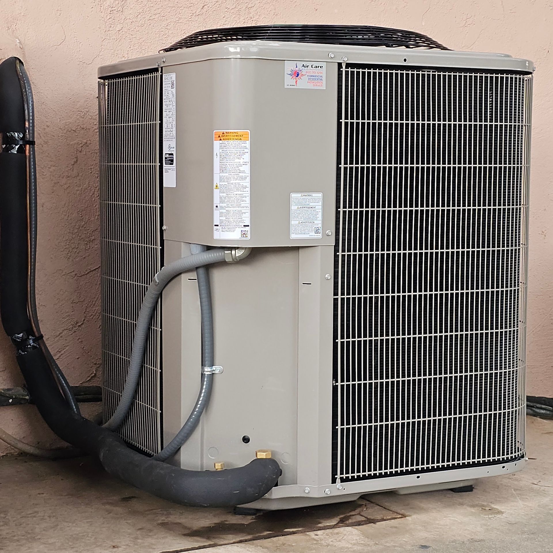 Outdoor air conditioner unit with gray metal casing and black hoses beside a wall