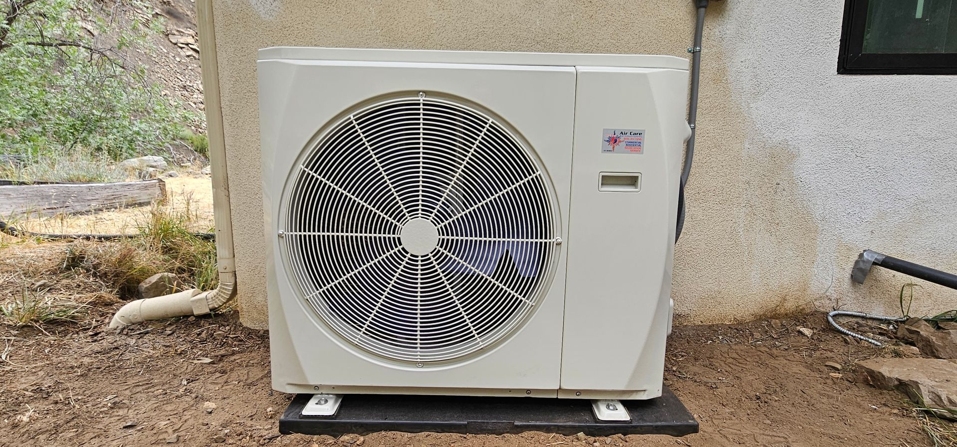 Outdoor HVAC unit beside a house wall on gravel, with a large front fan and white casing