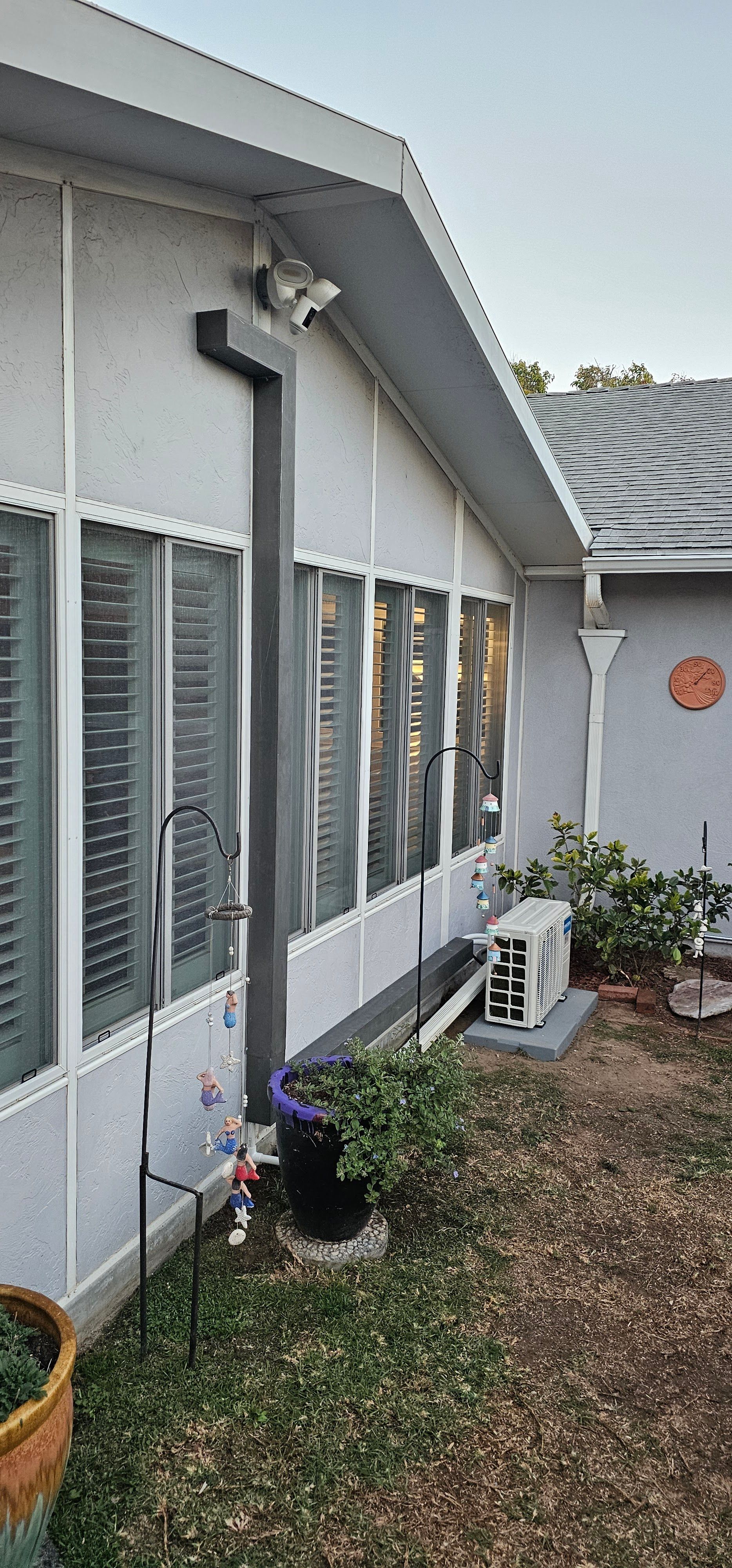 Side of a white house with windows, a wall-mounted light, and small potted plants along a gravel path.