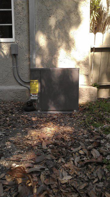Outdoor HVAC unit beside a house wall, surrounded by dry leaves and a fence gate.
