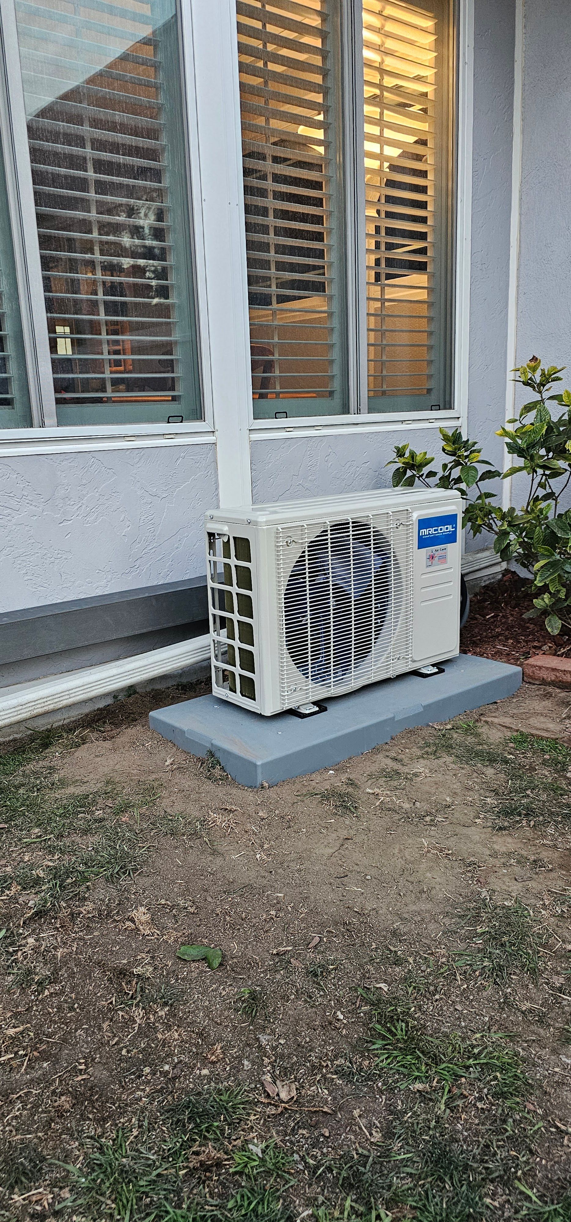 Outdoor air conditioner unit beside a house wall with shuttered windows and gravel ground