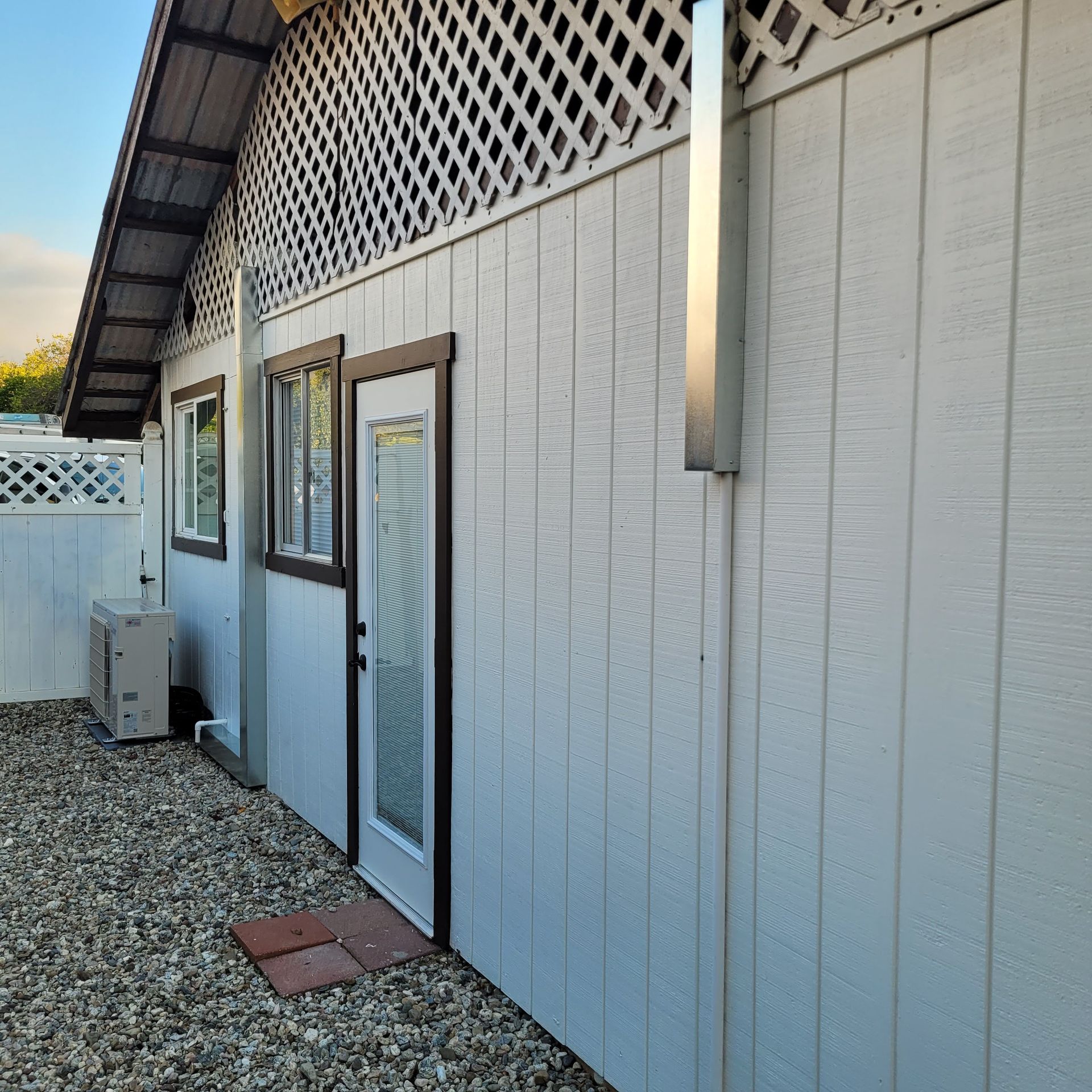 Side view of a white building with windows, a glass door, gravel ground, and a small HVAC unit nearby