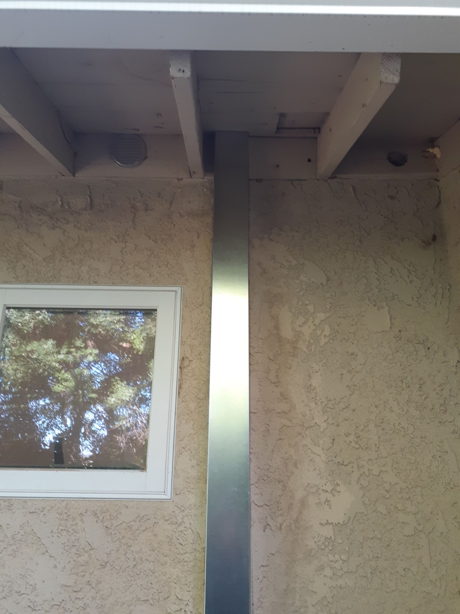 Vertical metal downspout on tan stucco wall beneath roof eaves, beside a small window with tree reflections