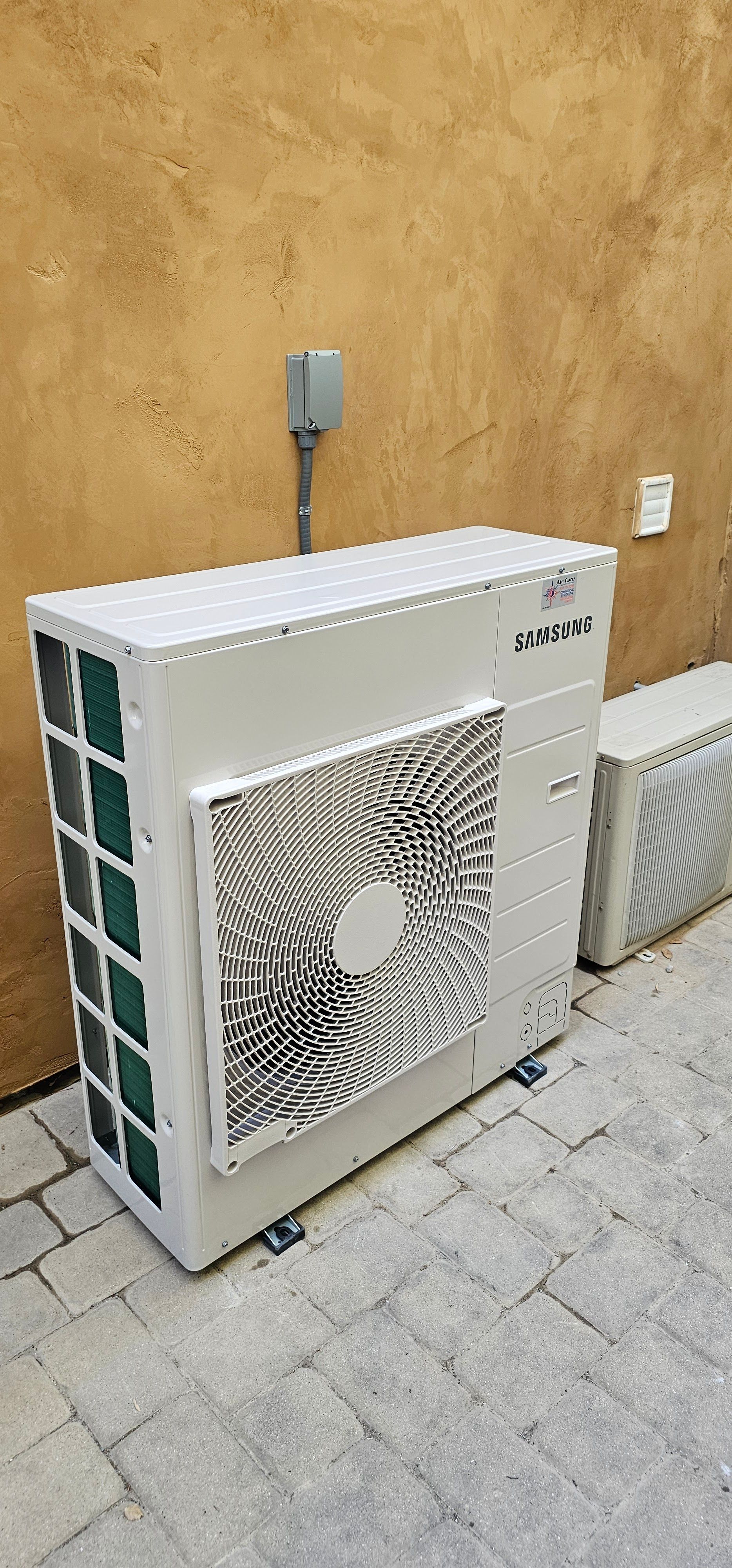 Outdoor air conditioning units beside a tan wall on a paved patio
