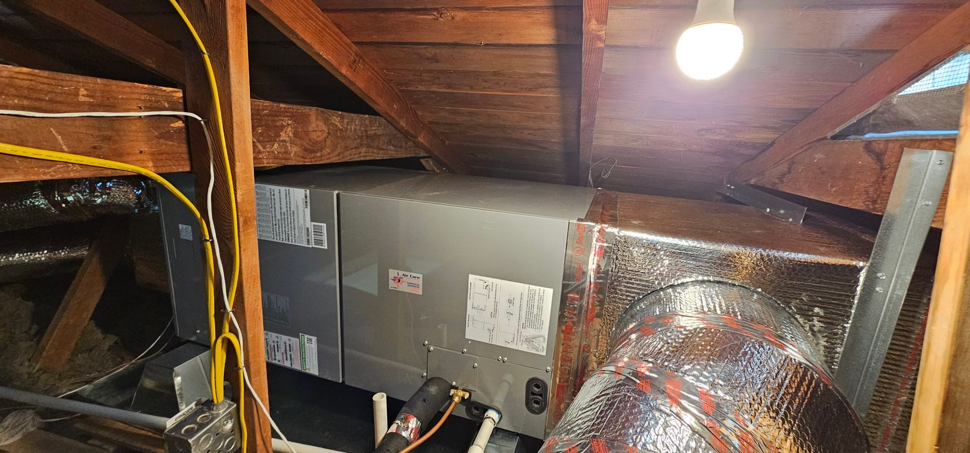 Attic with exposed ducts, insulation, and wiring under a wooden roof, lit by a single bulb