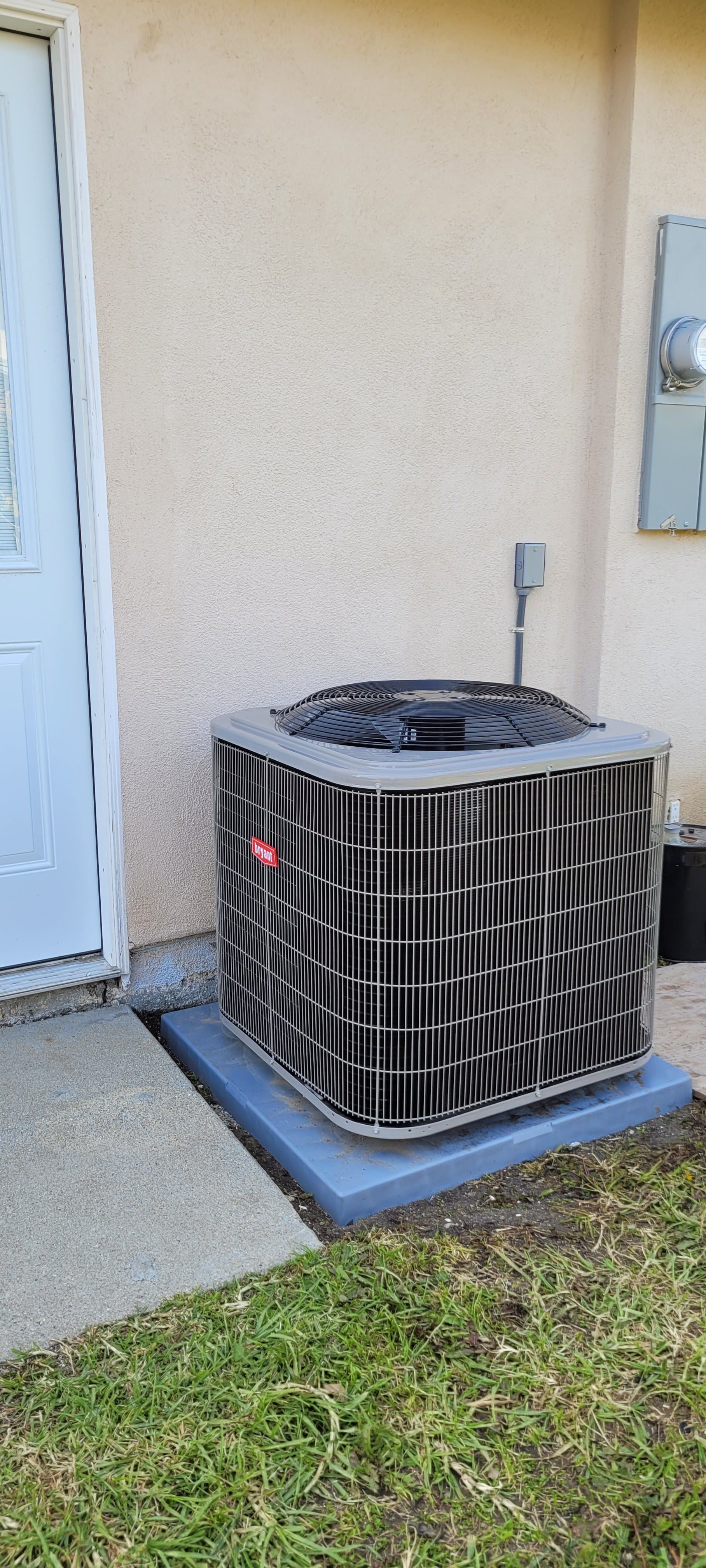 Outdoor air conditioner unit beside a house wall and door on a concrete pad.