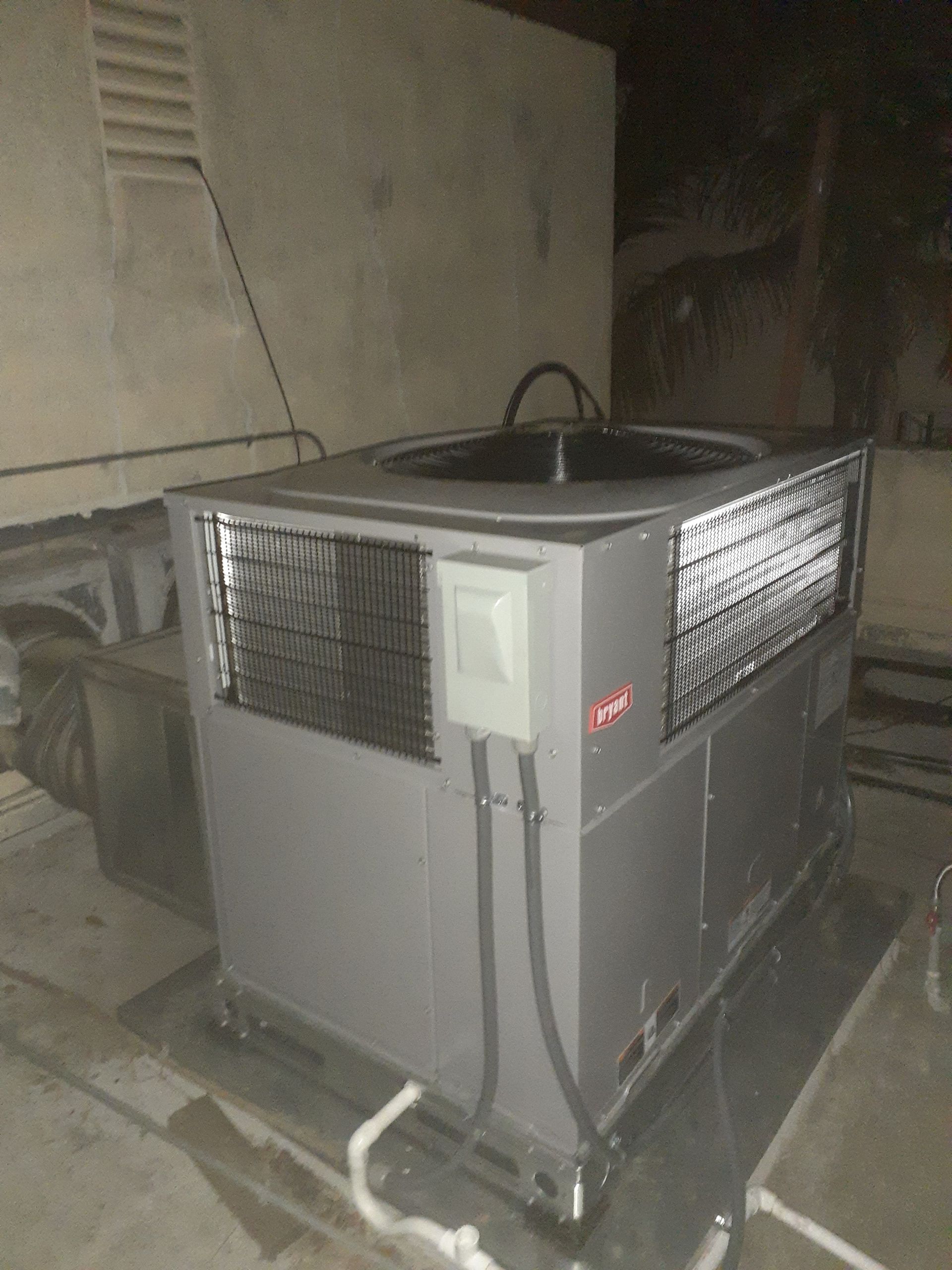 Outdoor HVAC unit on a concrete pad, with metal grilles and pipes beside a wall at night