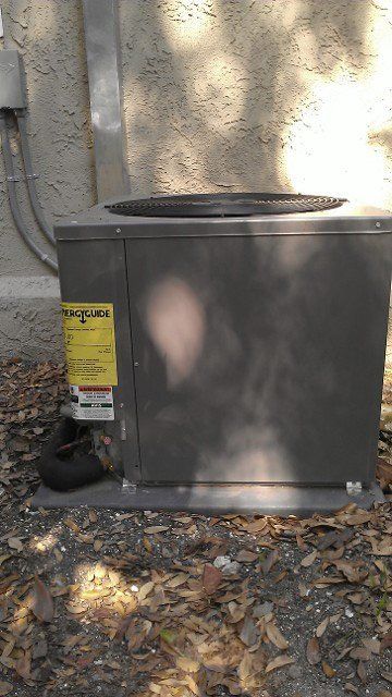 Outdoor gray HVAC unit beside a wall, with a yellow label and fallen leaves on the ground