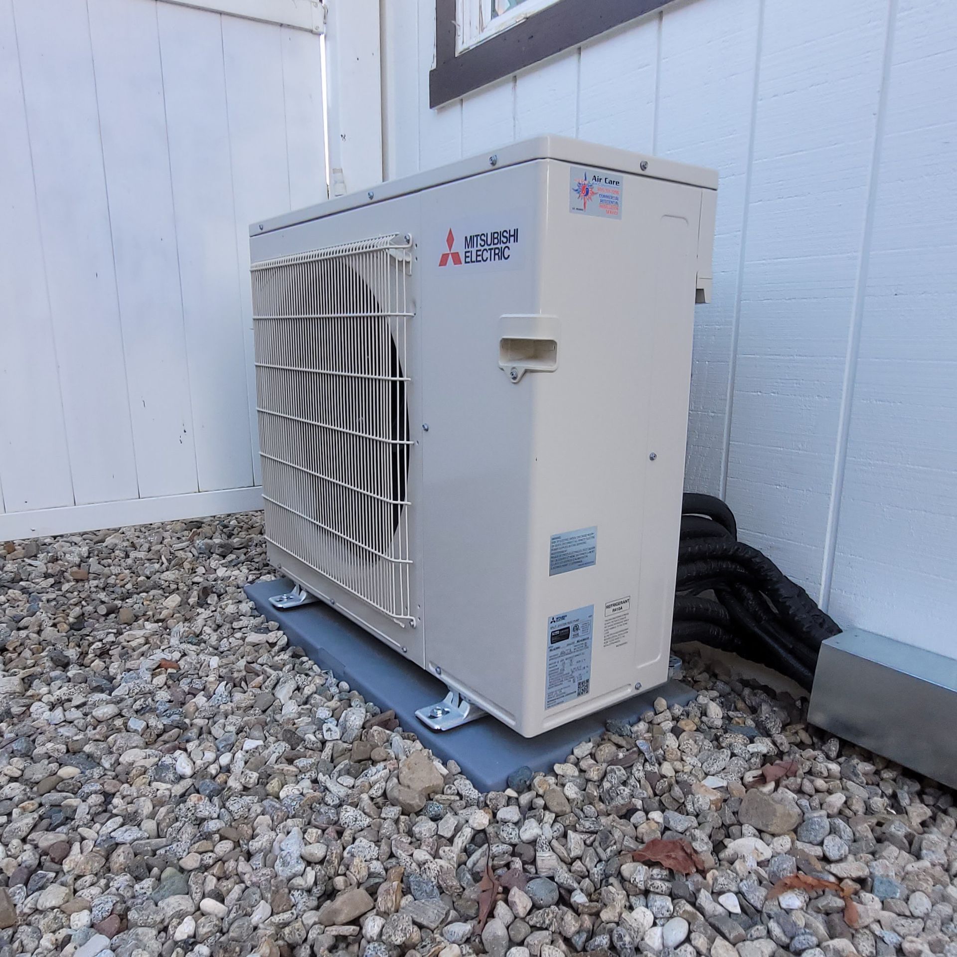 White outdoor HVAC unit on gravel beside a building wall, with black hoses behind it