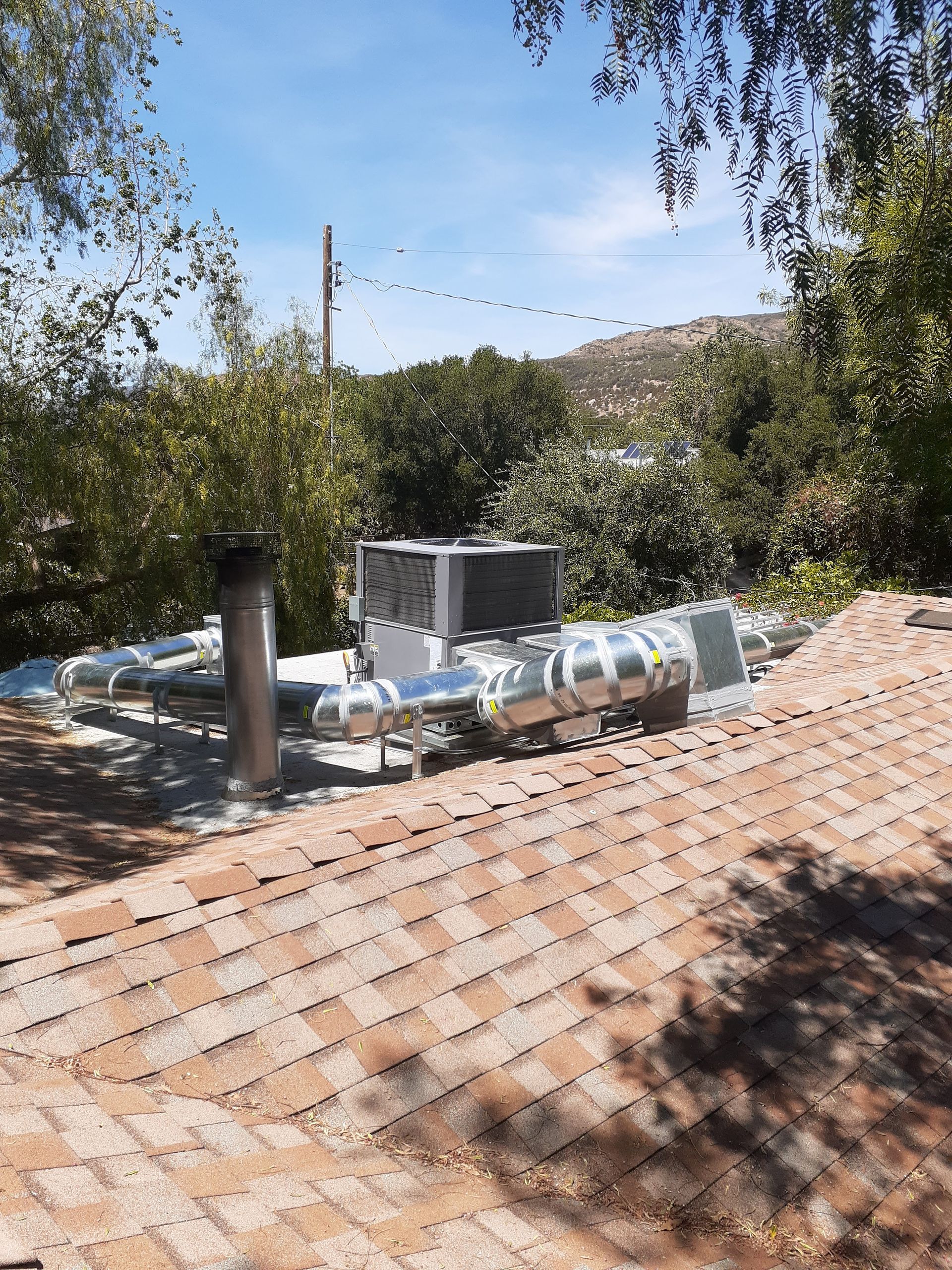 Rooftop HVAC unit and metal vents on a tiled roof, framed by trees and a blue sky