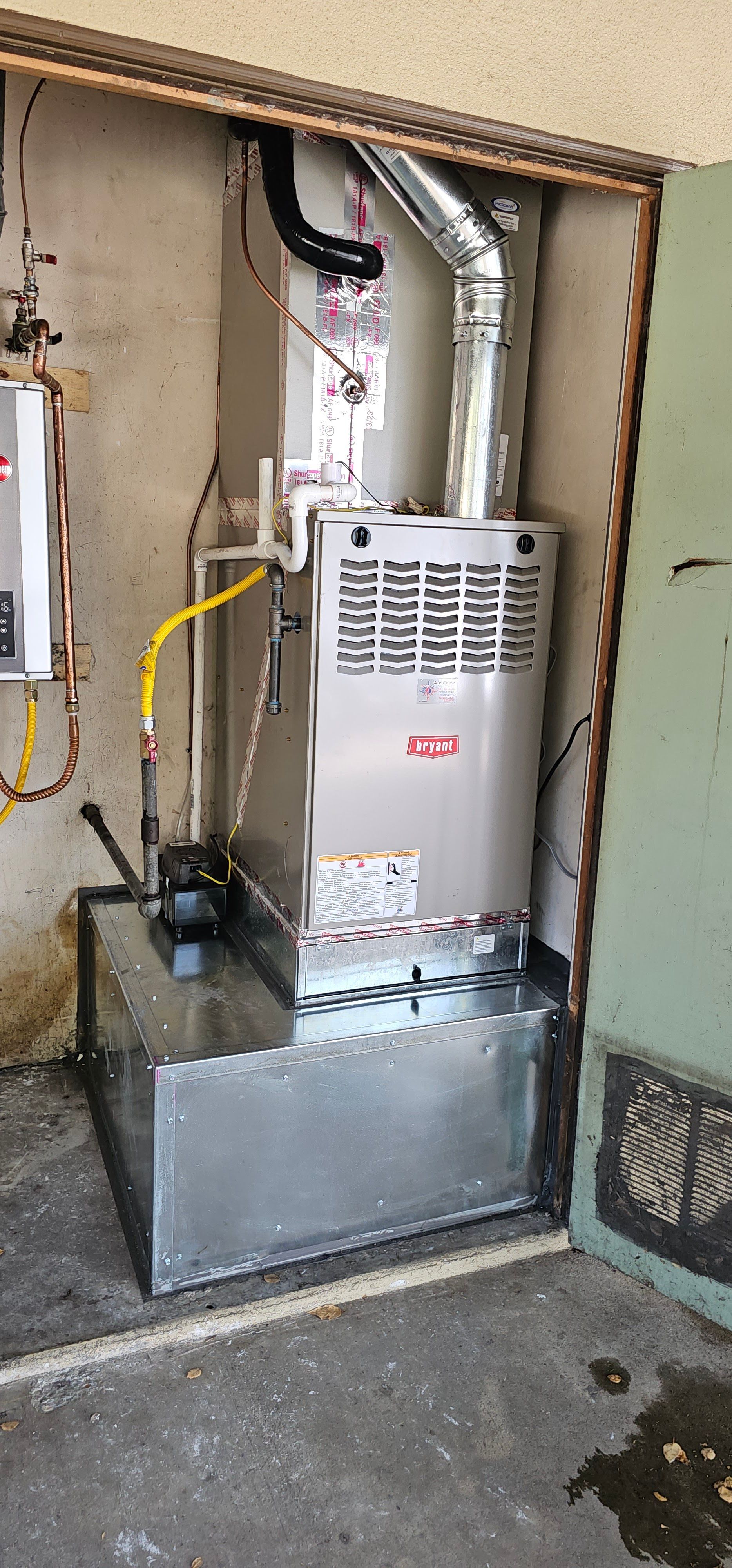Old furnace in a small utility alcove with exposed pipes and a metal duct, beside a concrete floor.