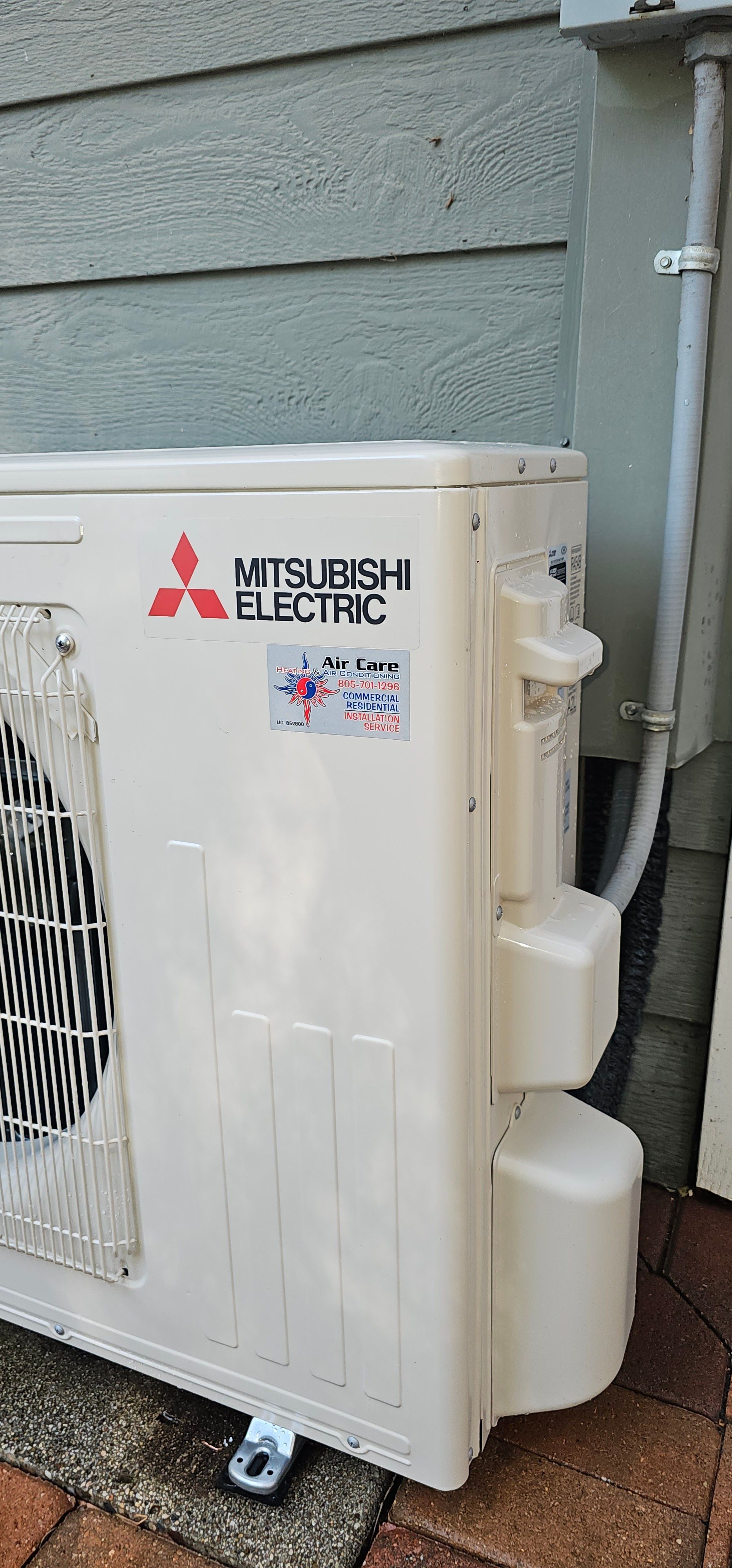 White Mitsubishi Electric outdoor heat pump unit mounted beside a gray house wall.
